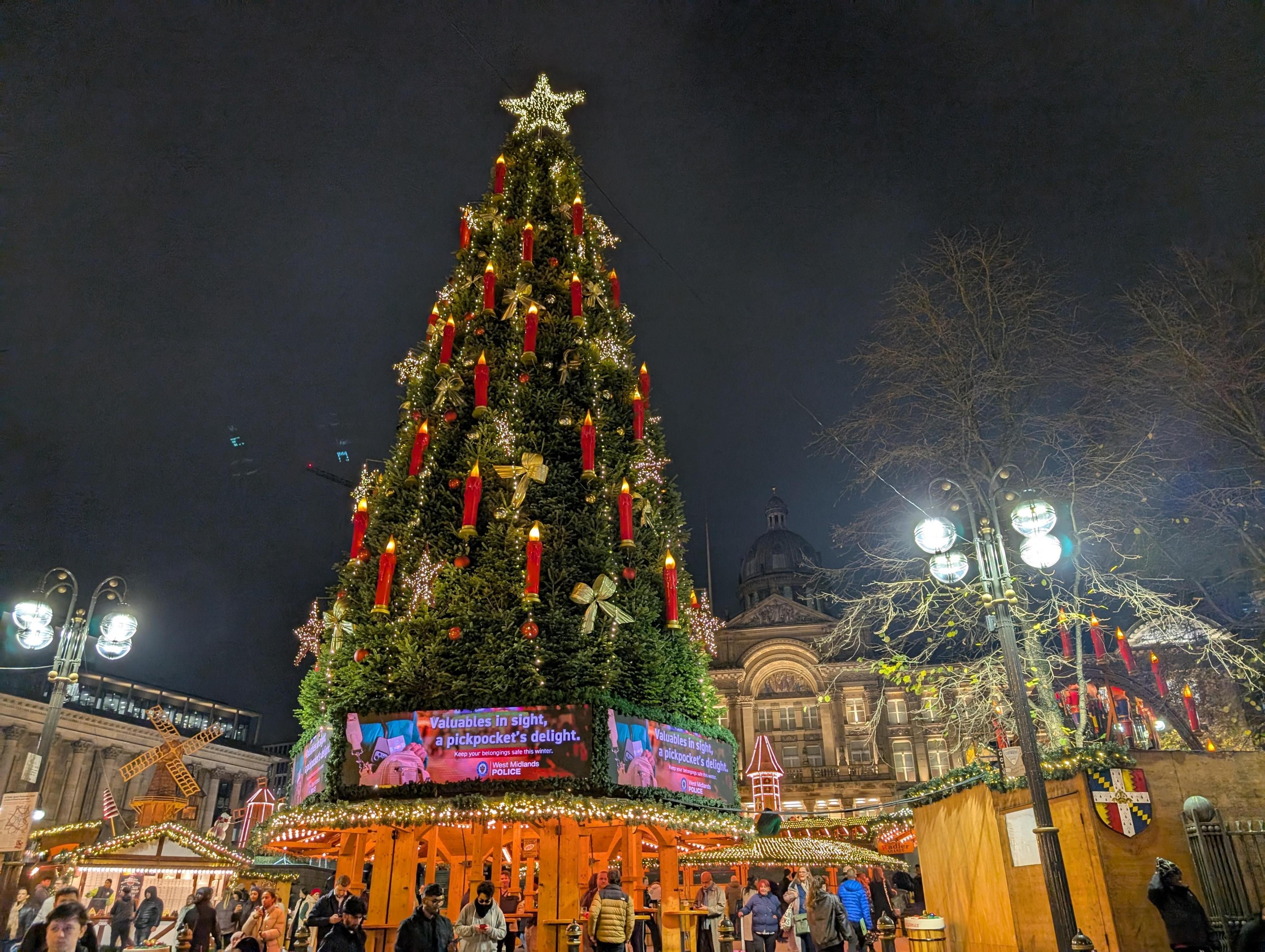 A large, ornate Christmas tree sits above a wooden structure where people are sitting to eat and drink. Around the base are screens showing police warnings about the risk of pickpocketing.