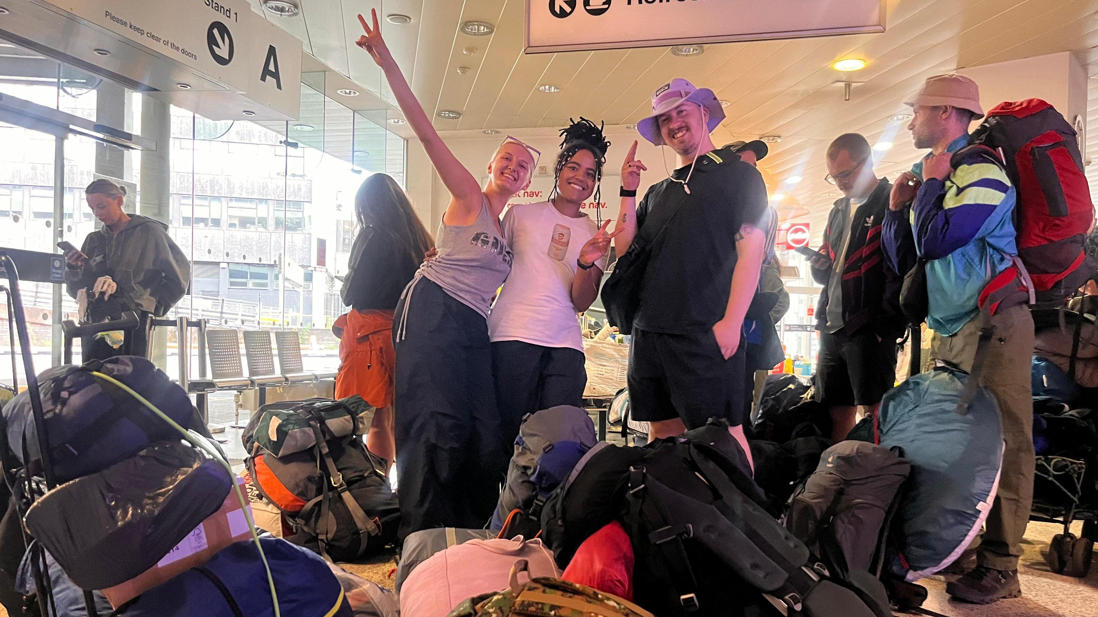 Laurence wears a black T-shirt, black shorts and a purple hat. He stands next to his friends Meg (left) and Kitty (middle) in the coach station surrounded by bags. They smile at the camera as Meg and Kitty do peace signs