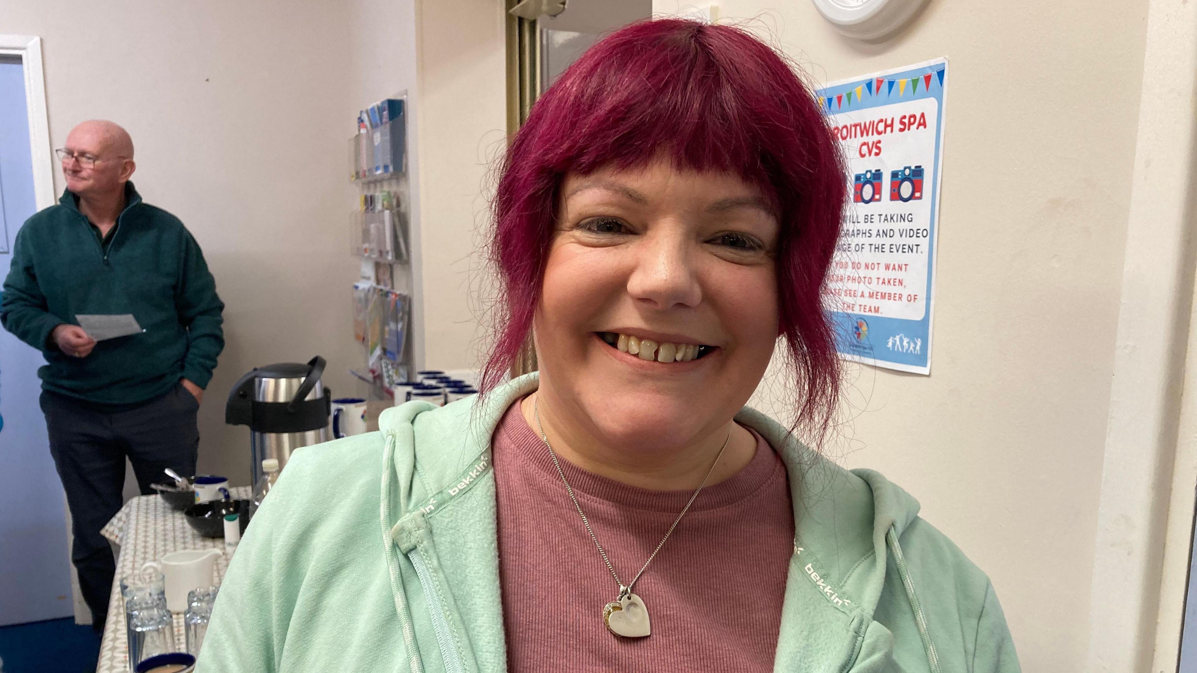 A woman with dyed red hair is smiling at the camera. She is wearing a mauve sweater, a mint green hoodie and a heart necklace. There is a table with a large coffee flask and glasses behind her.