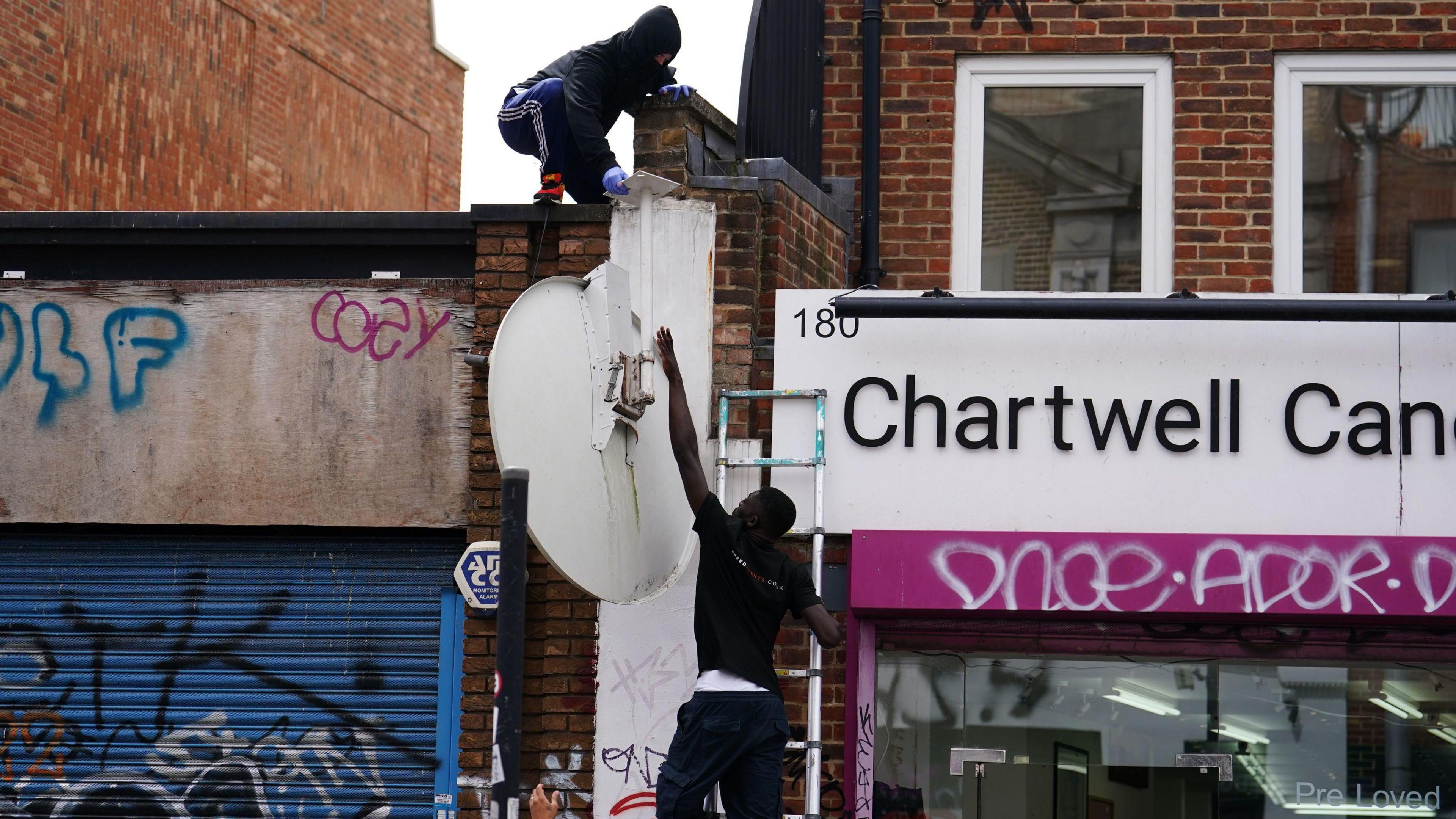 Satellite dish featuring the howling wolf stencil being handed down from person wearing a balaclava to another man, wearing black t-shirt and is also steadying a metal ladder. In the background are the shopfronts, which have graffiti on them.