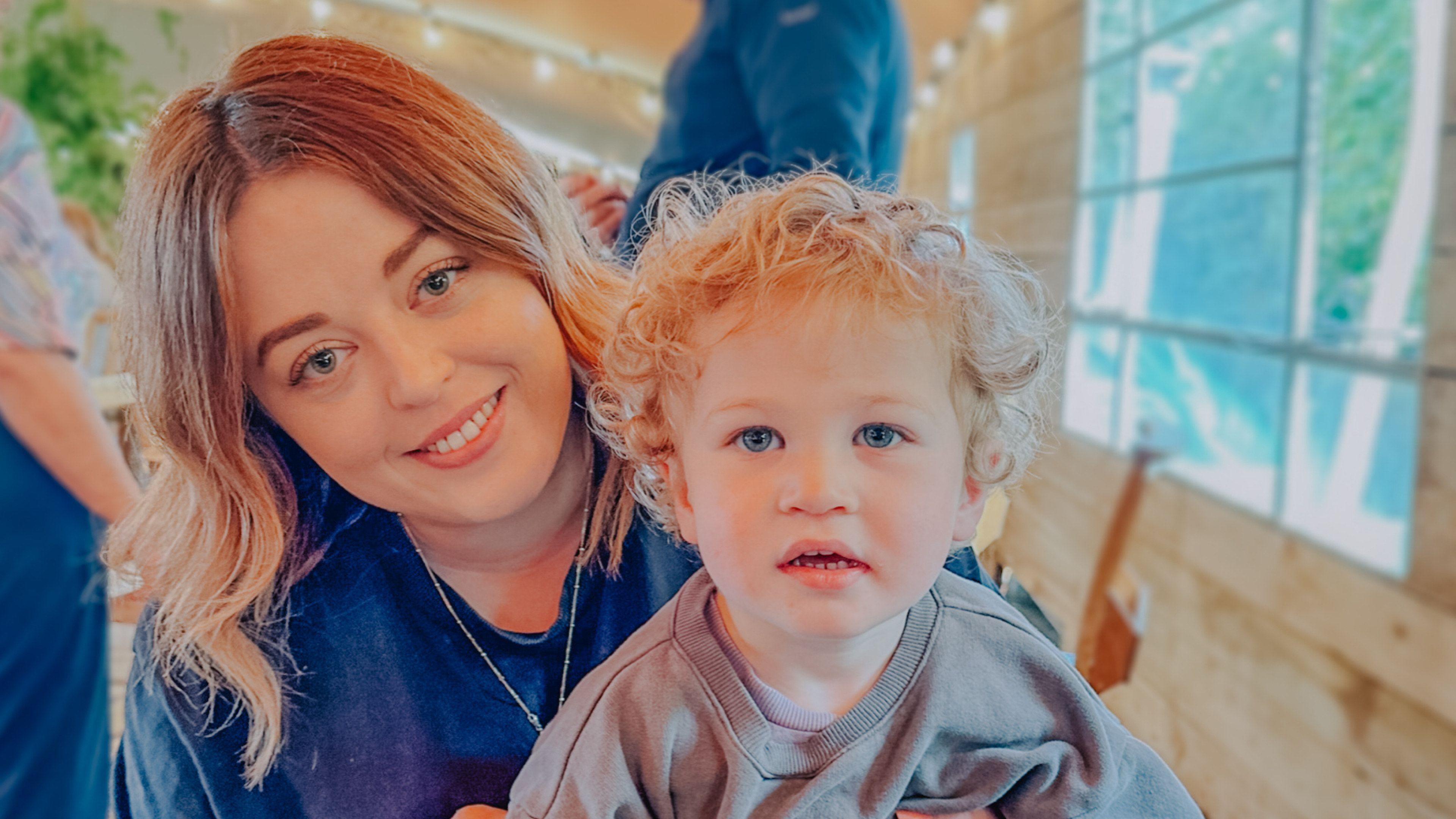 A woman with curly blonde hair smiles at the camera with her little boy who has blonde short curly hair