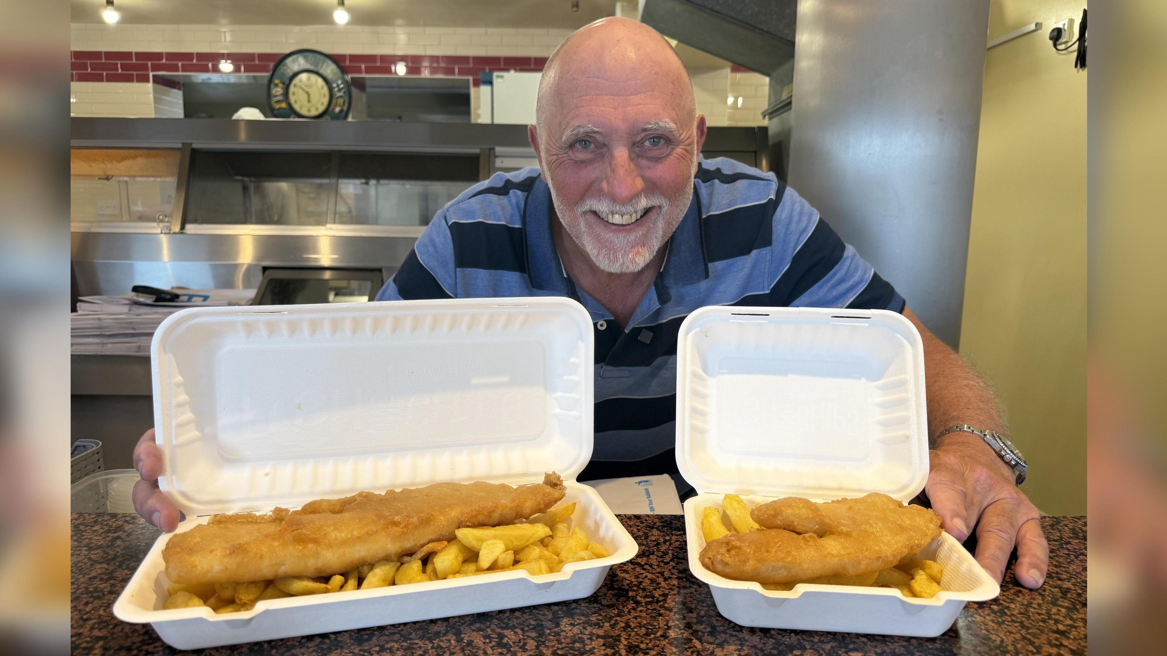 A man with a bald head, smiling, holdding two portions of fish and chips.