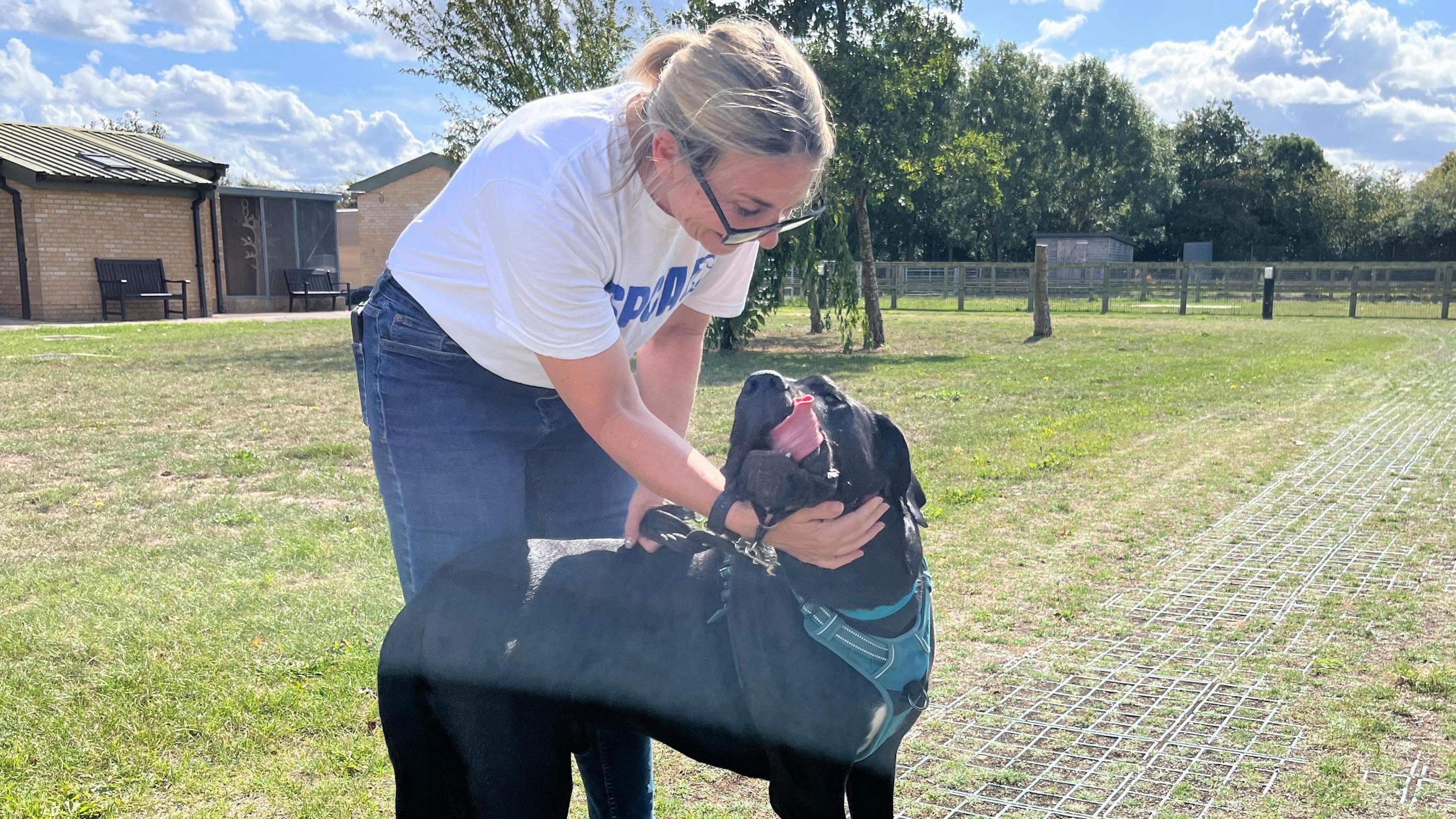 Nicola Walker standing outside. She is wearing a white T-shirt with a blue RSPCA logo and writing on it. She is crouched down petting a black dog which has it's tongue sticking out.