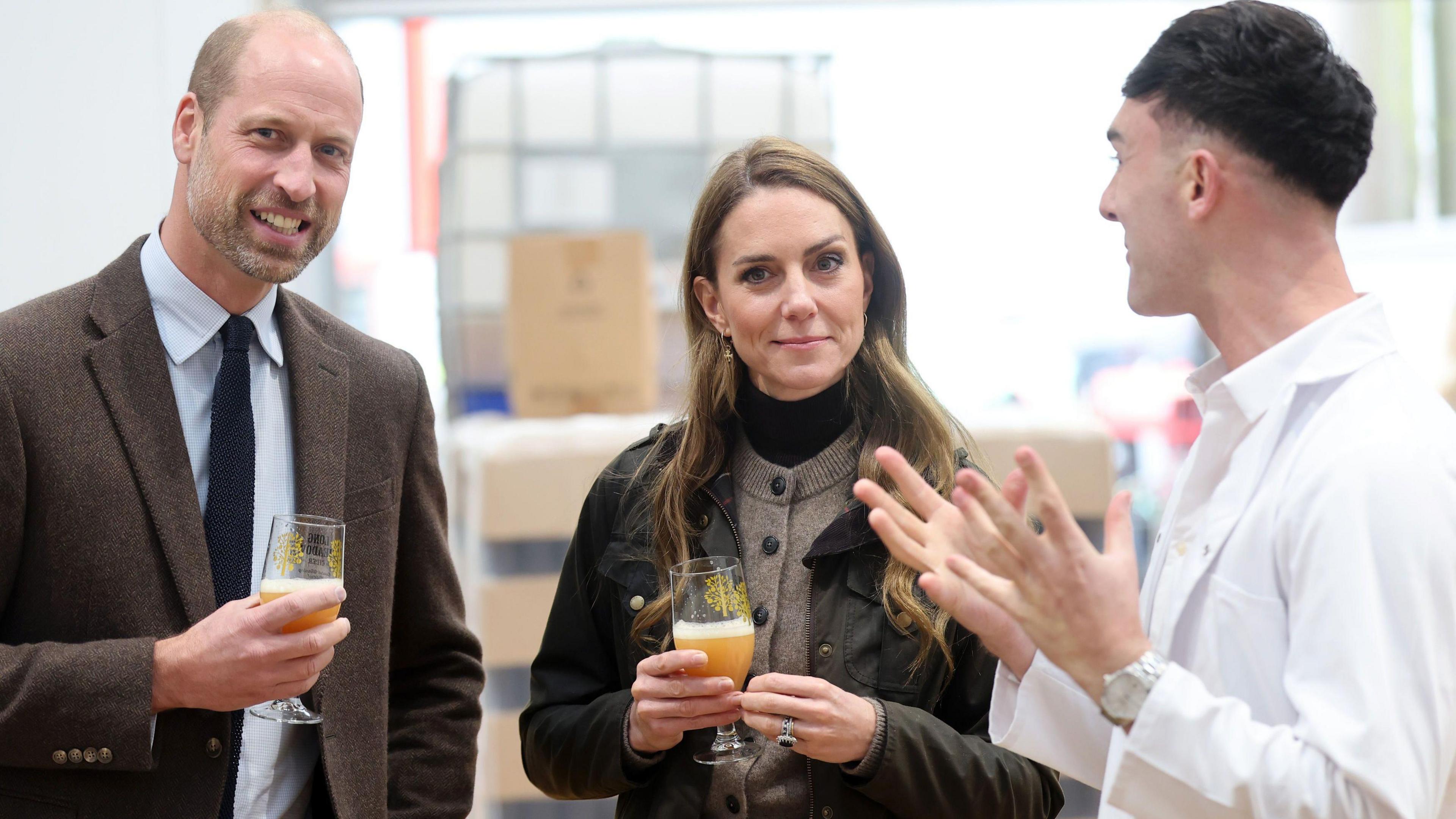 The prince and princess look into the camera whilst a man with short black hair wearing a lab coat stands side on conversing with them. William who is on the far left of the frame wears a brown blazer, blue shirt and navy tie, whilst holding a half full glass of a fizzy cloudy-orange liquid. Catherine, who holds an identical glass of the same liquid, stands in the middle wearing a green coat, brown waistcoat and black turtle neck sweater. They are inside a factory with white walls.
