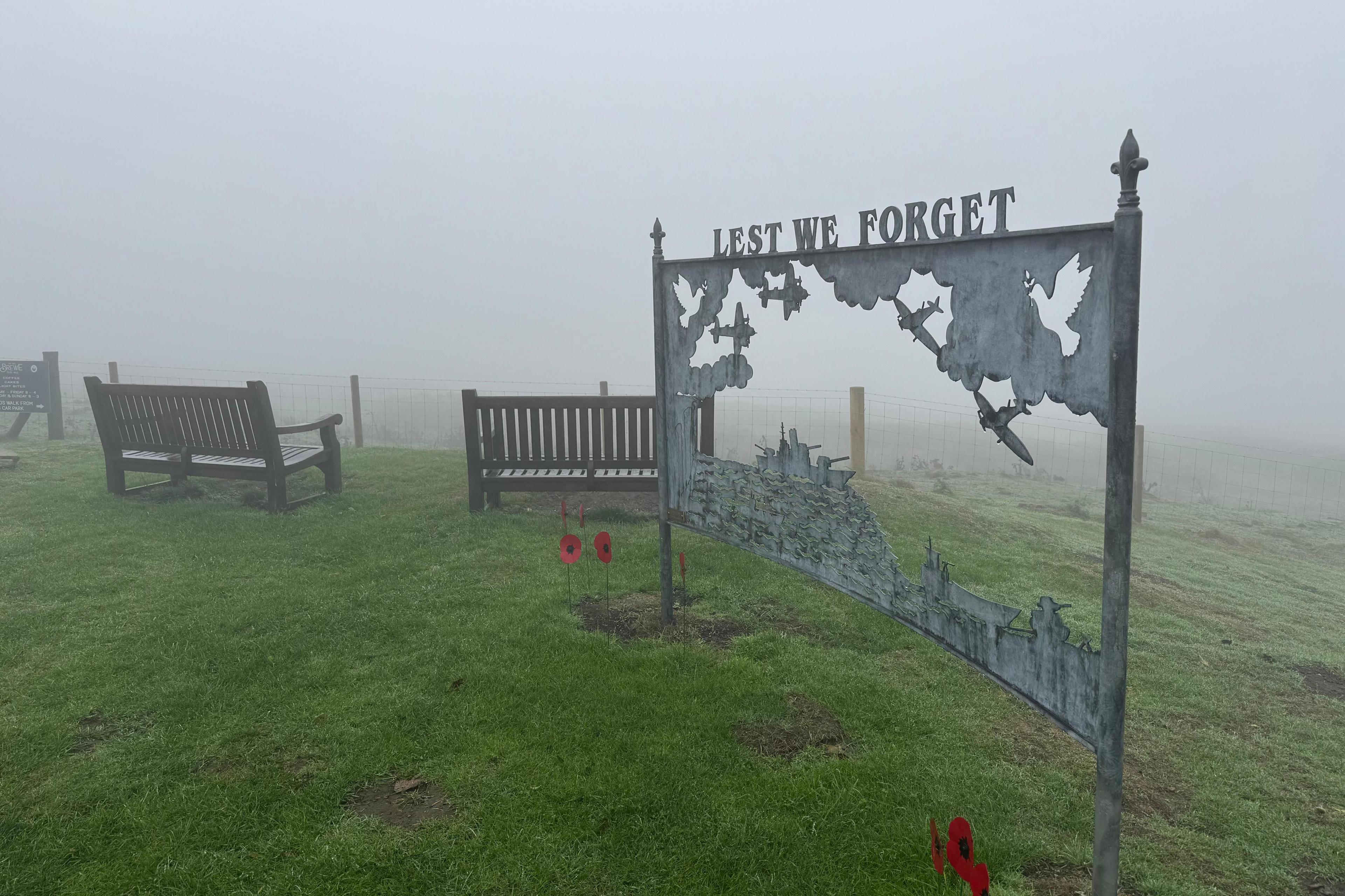 A tableau in grey, weathered metal featuring a cut-out scene of World War Two aircraft among clouds at the top with naval ships at sea underneath. The posts supporting the scene are topped with fleur-de-lys finials, with cut-out lettering reading "Lest we forget" across the top. 