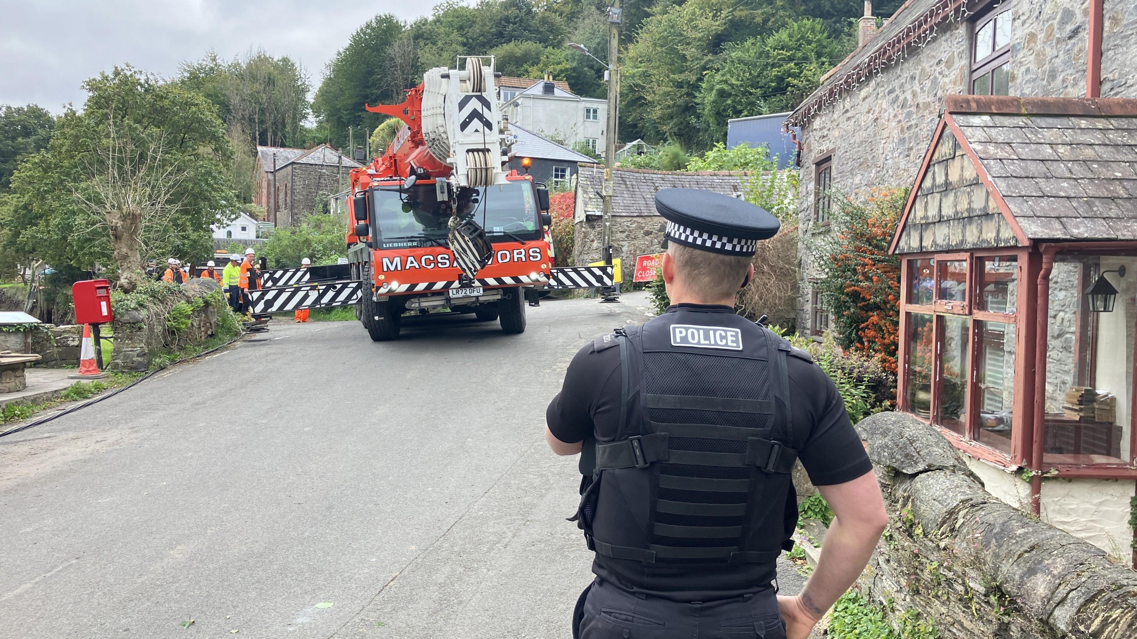 A black, red and white crane on a road beside a stone house on a village, with a policeman in the foreground.