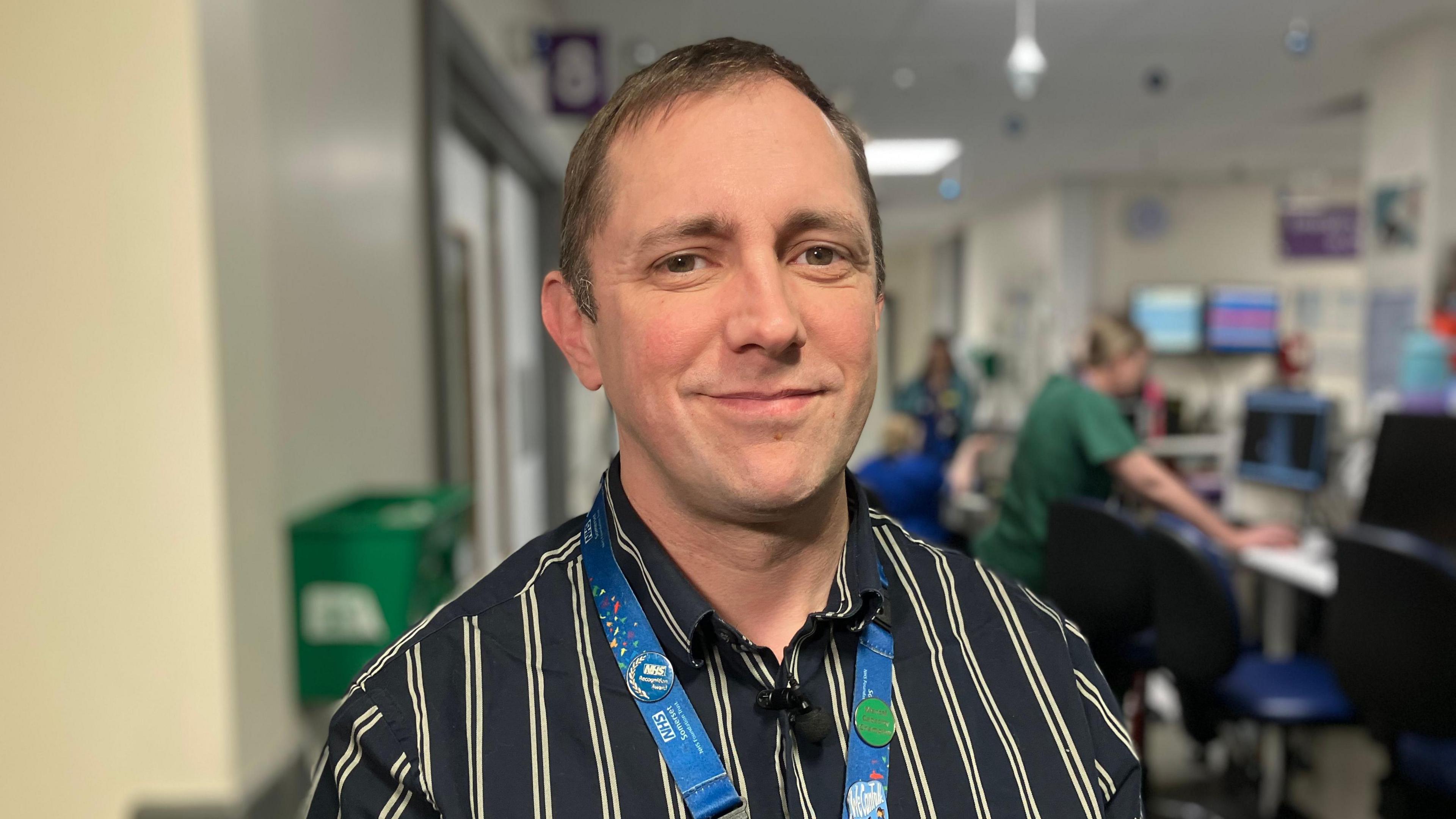 A man with short brown hair smiling into camera. He is wearing a black shirt with white stripes and a blue ID lanyard. Behind him are nurses at workstations in an A&E.