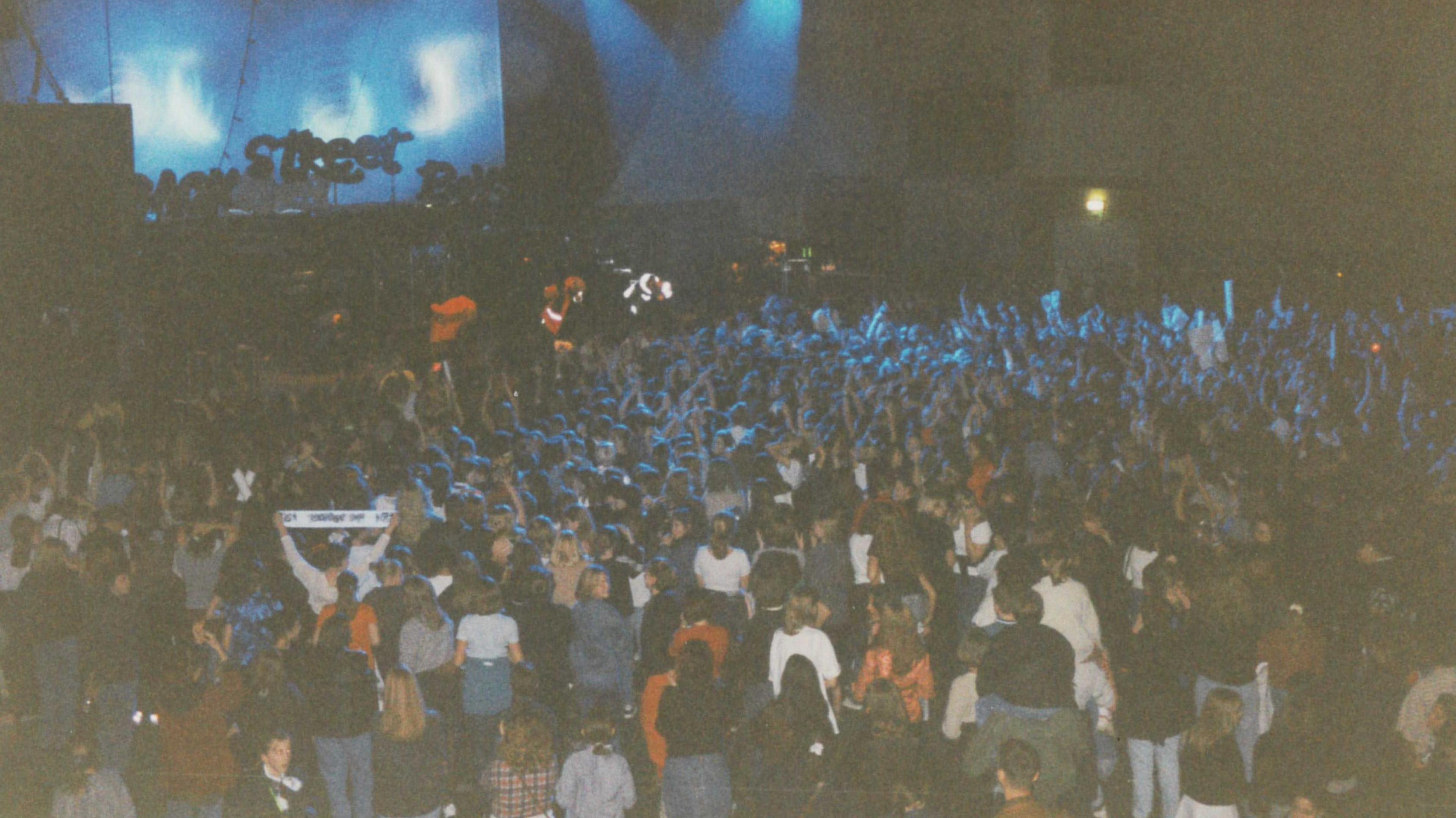 A shot from the back of the room showing the Backstreet Boys onstage and a large crowd in the toom. Some are holding up scarves and the photo is very grainy and 90s-looking