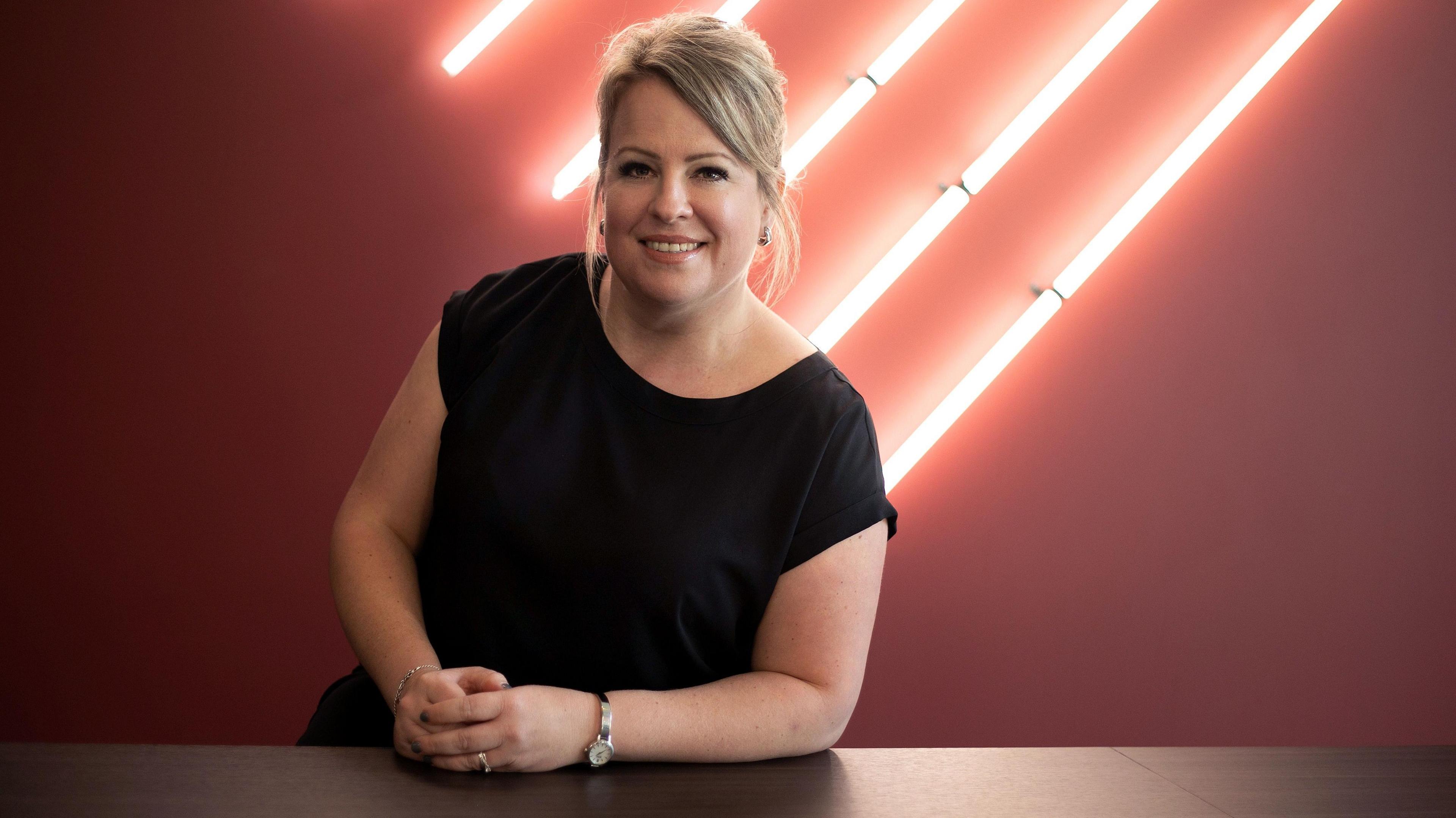 Sharon Livermore, looking at the camera, smiling, wearing a black outside, in front of a backdrop that shows a number of neon light strips 