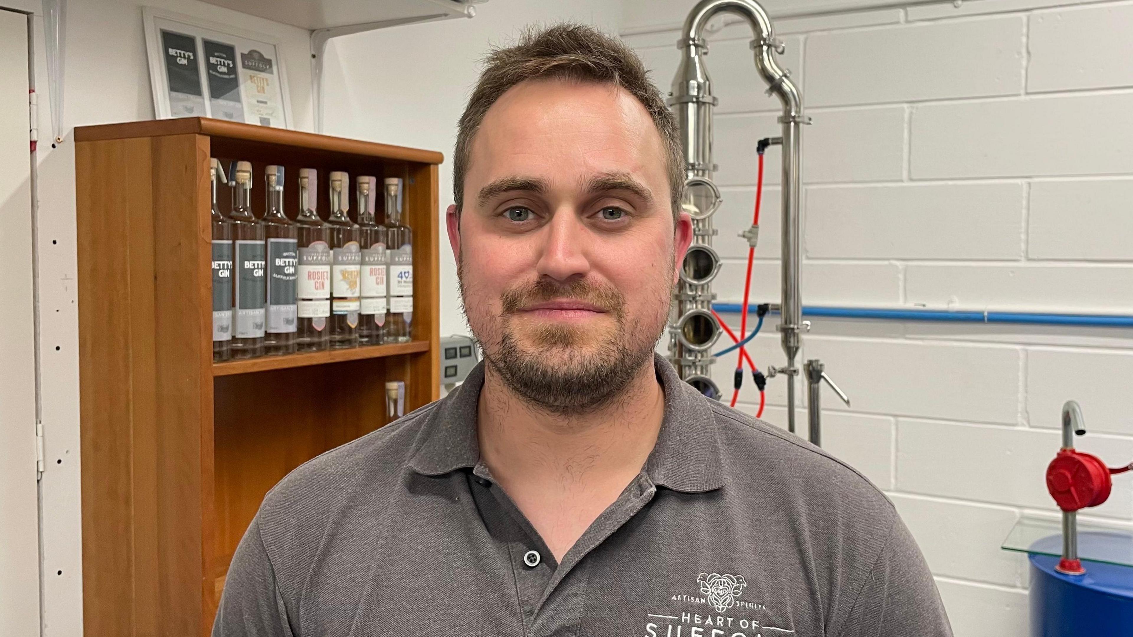 Ryan Luke is looking directly at the camera. He is wearing a dark grey, collared T-shirt, which is unbuttoned. Part of a Heart of Suffolk Distillery logo is visible on the chest. In the background are bottles of spirits on a shelf and some distilling equipment. Ryan has a stubbly brown beard, and short brown hair and blue eyes.