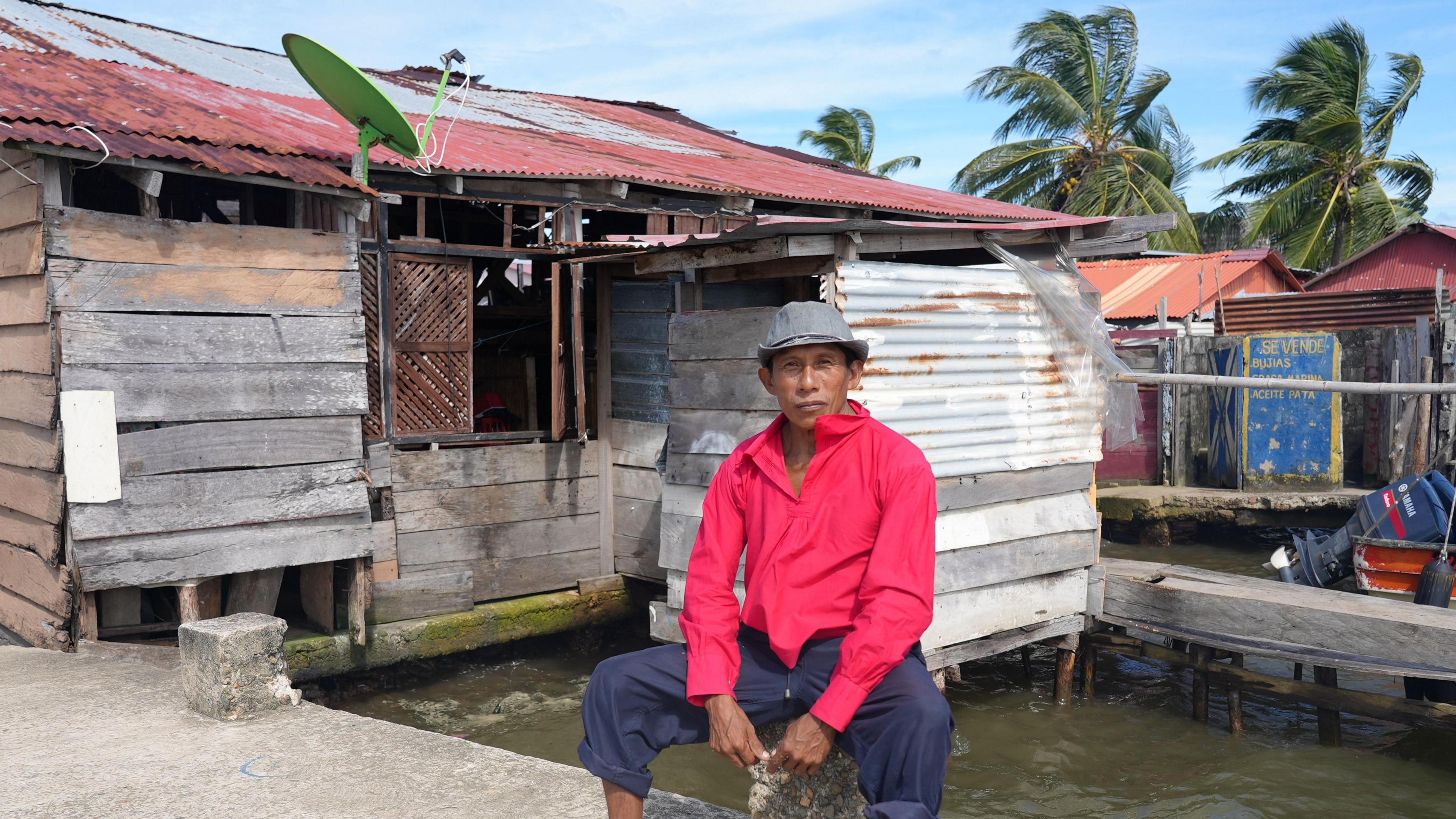 Delfino, in a bright pink shirt and grey hat sits on a low concrete post on a jetty, with a house built of wood and corrugated metal behind him. Part of the house is on stilts, sticking out into the water.