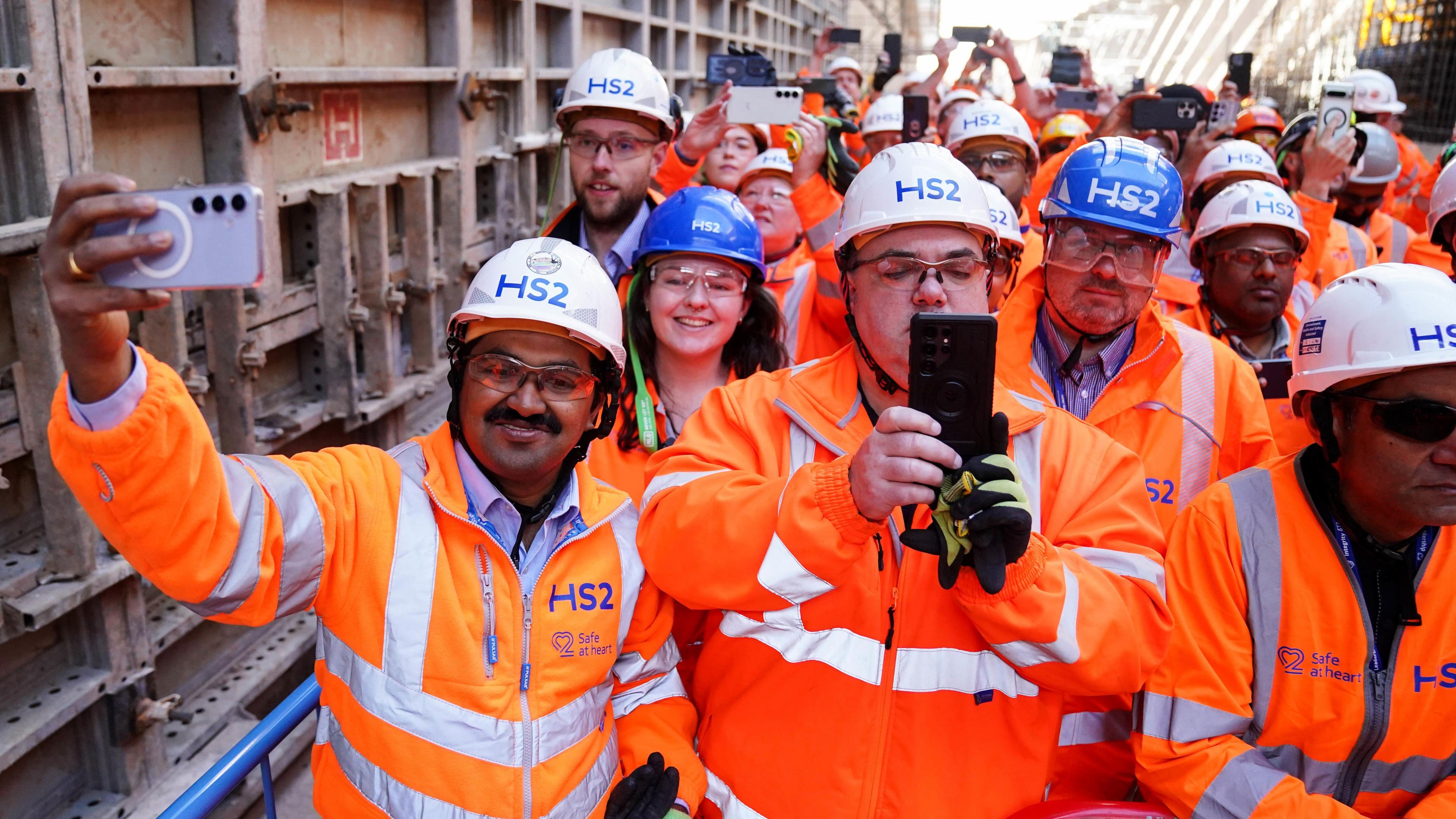 Men and women in orange high-vis jackets are standing with their phones out. They have white hard hats on that read "HS2" in blur writing. They are wearing protective goggles.