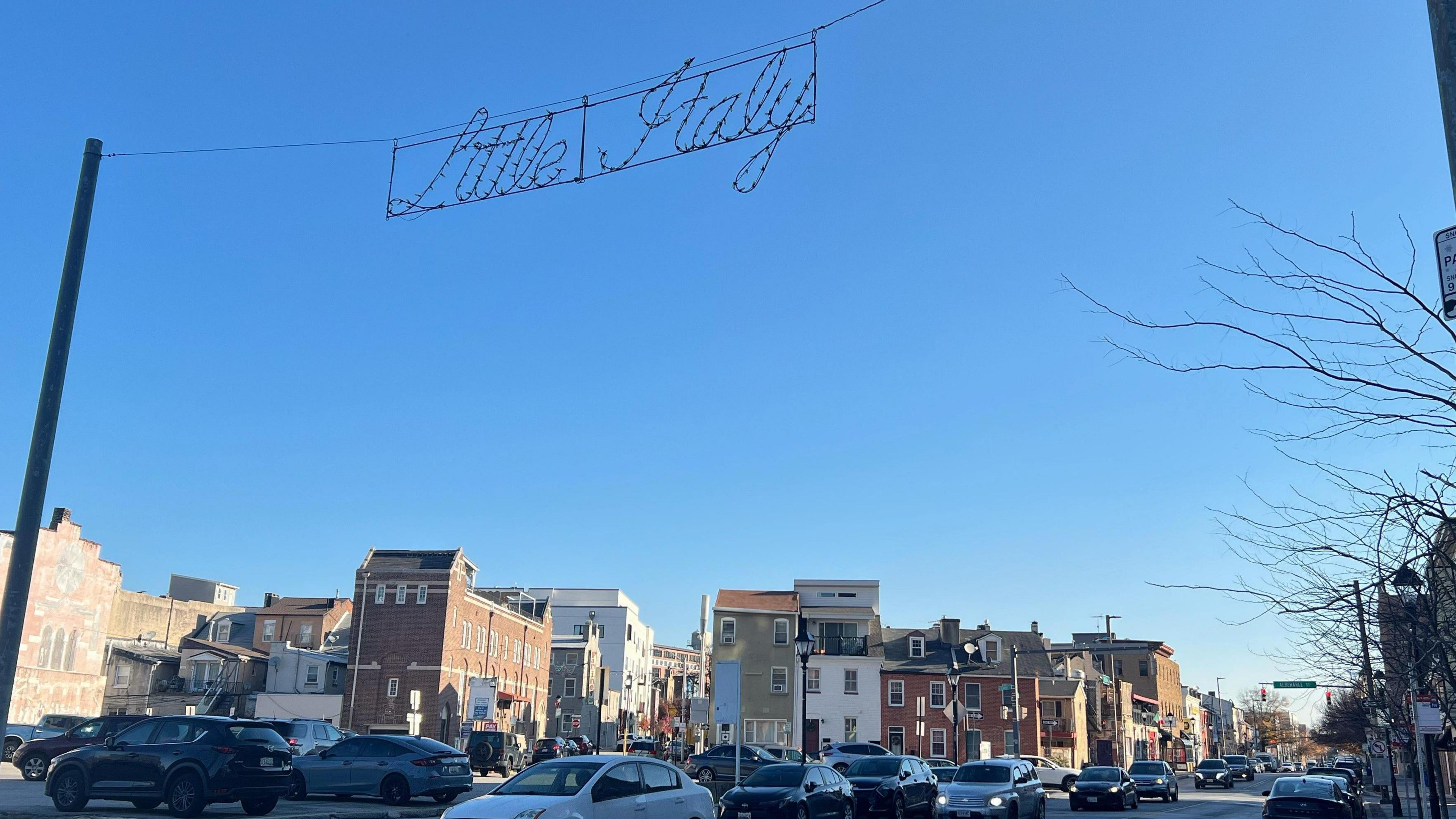 A lighted sign that says Little Italy is suspended over a street against a blue sky with brick and wood buildings behind it. There are cars driving below it on the street.