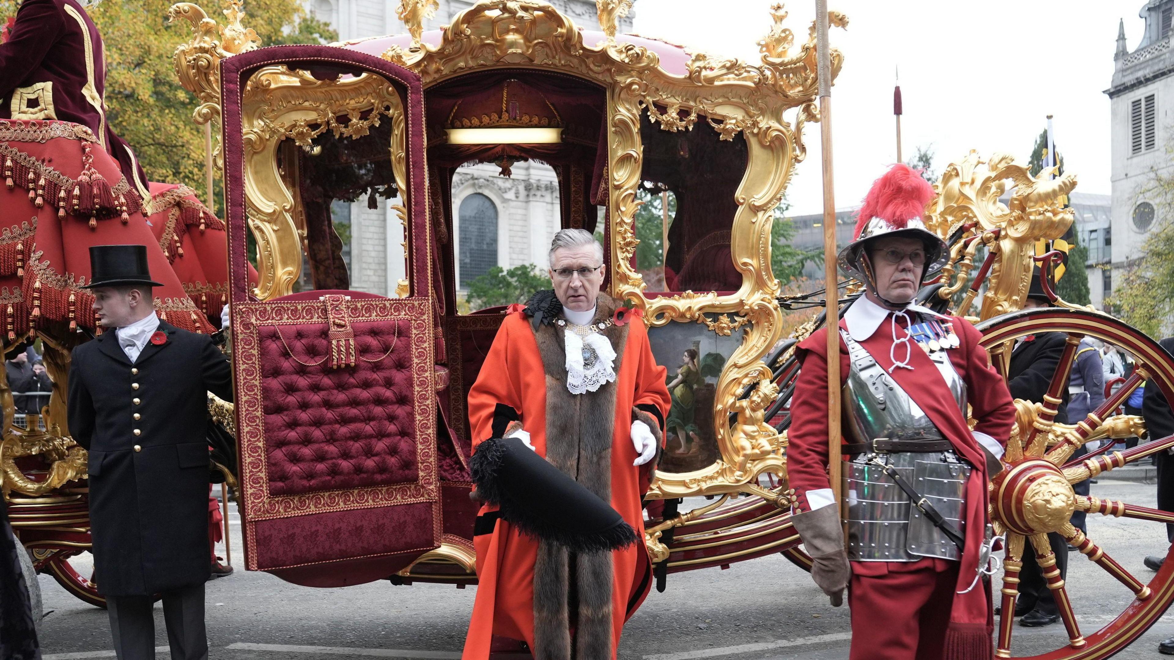 The Lord Mayor of London steps out of an ornate, gilded state carriage, dressed in traditional red robes with fur trim and carrying a ceremonial hat. Beside him stands a member of the ceremonial guard in historic armour and a red uniform, holding a pike. Another attendant in a black coat with a top hat holds the carriage door open. 