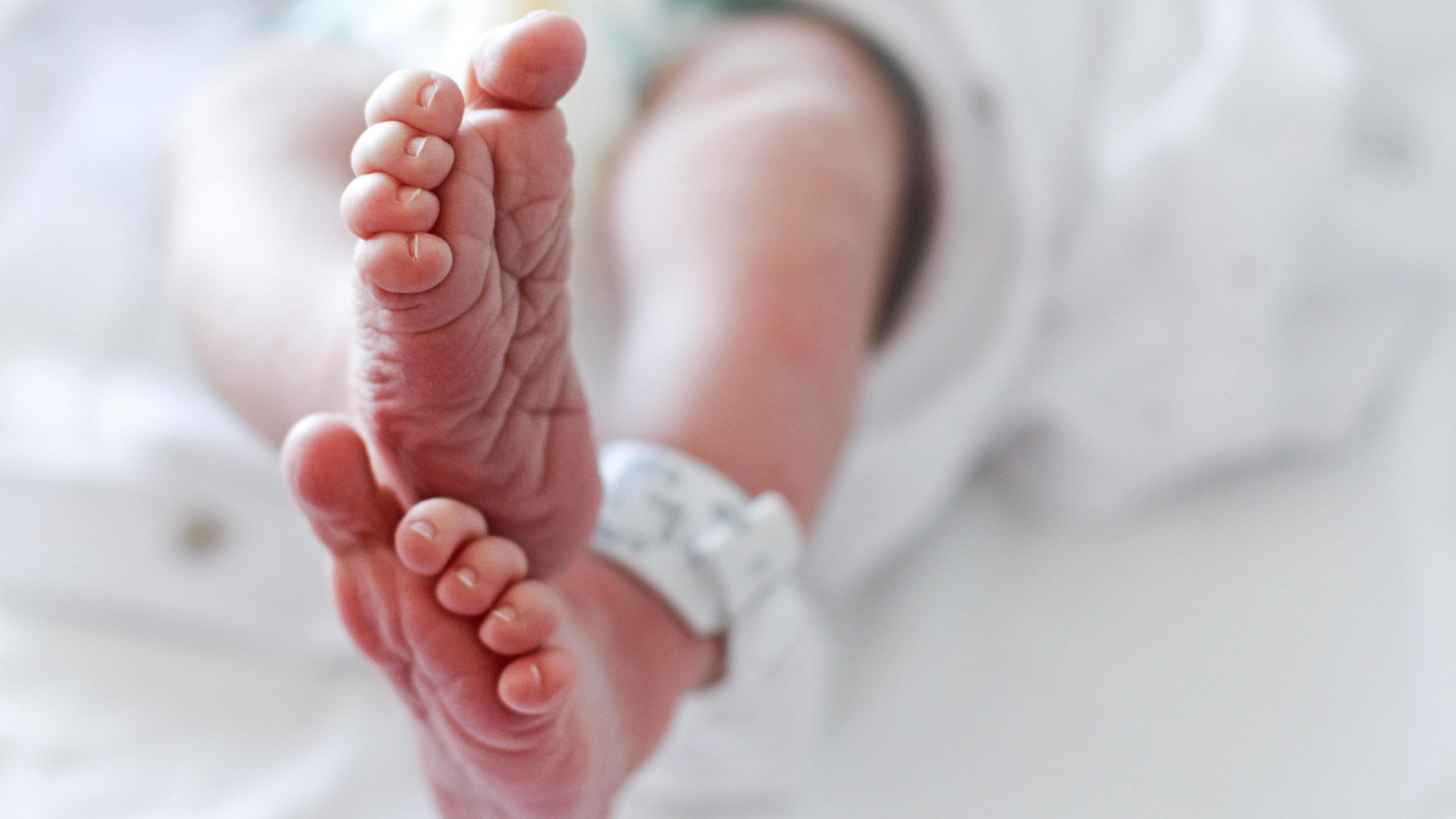 A newborn baby's feet with a hospital identity tag. The baby is wearing white, with white blankets and sheets are in the background.