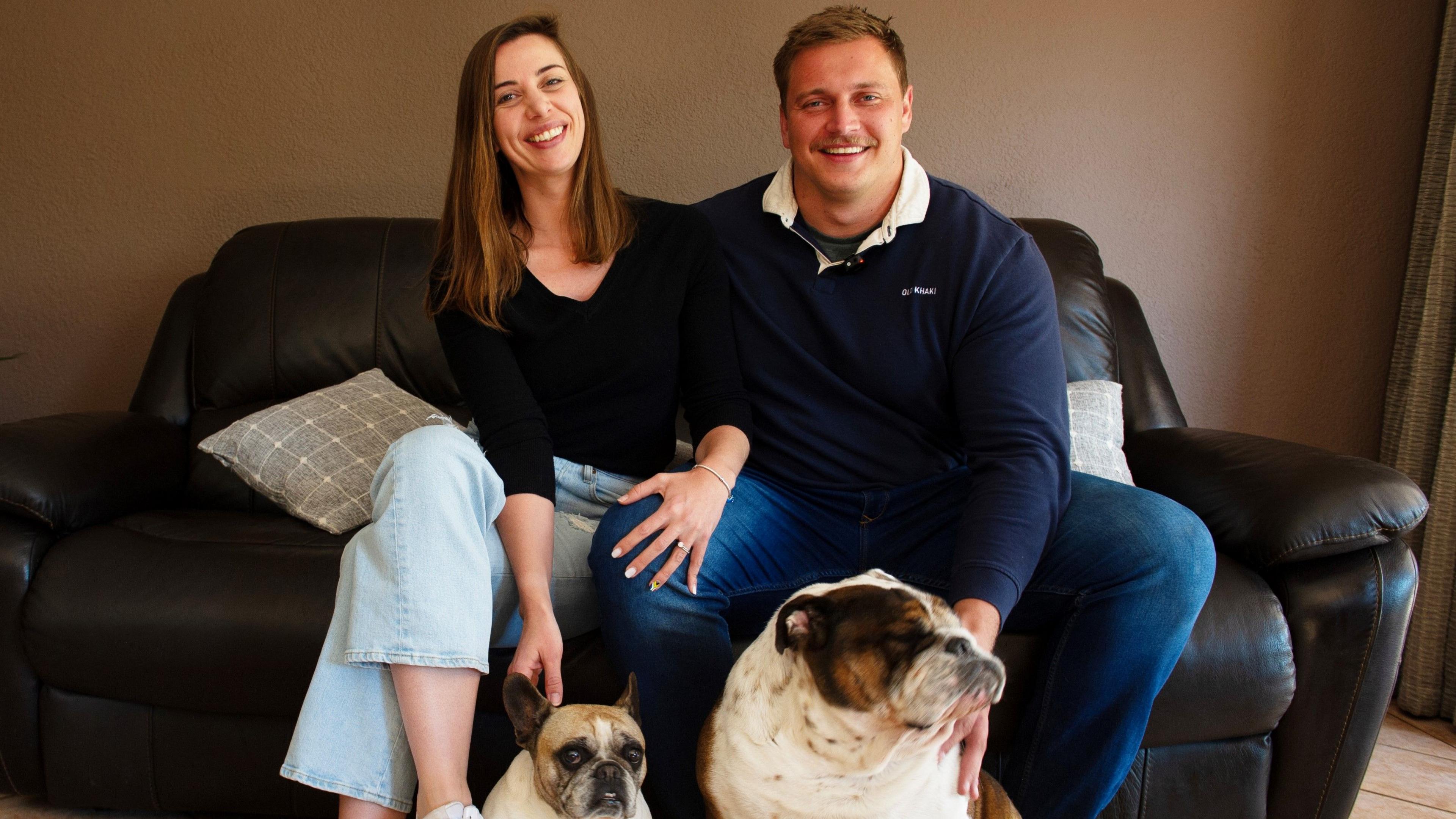 An at-home image of Rayno Nel and his wife Monja sitting next to each other on a dark leather sofa and smiling into the camera. Monja has her left hand resting on Nel's knee, while in front of them sit their two dogs
