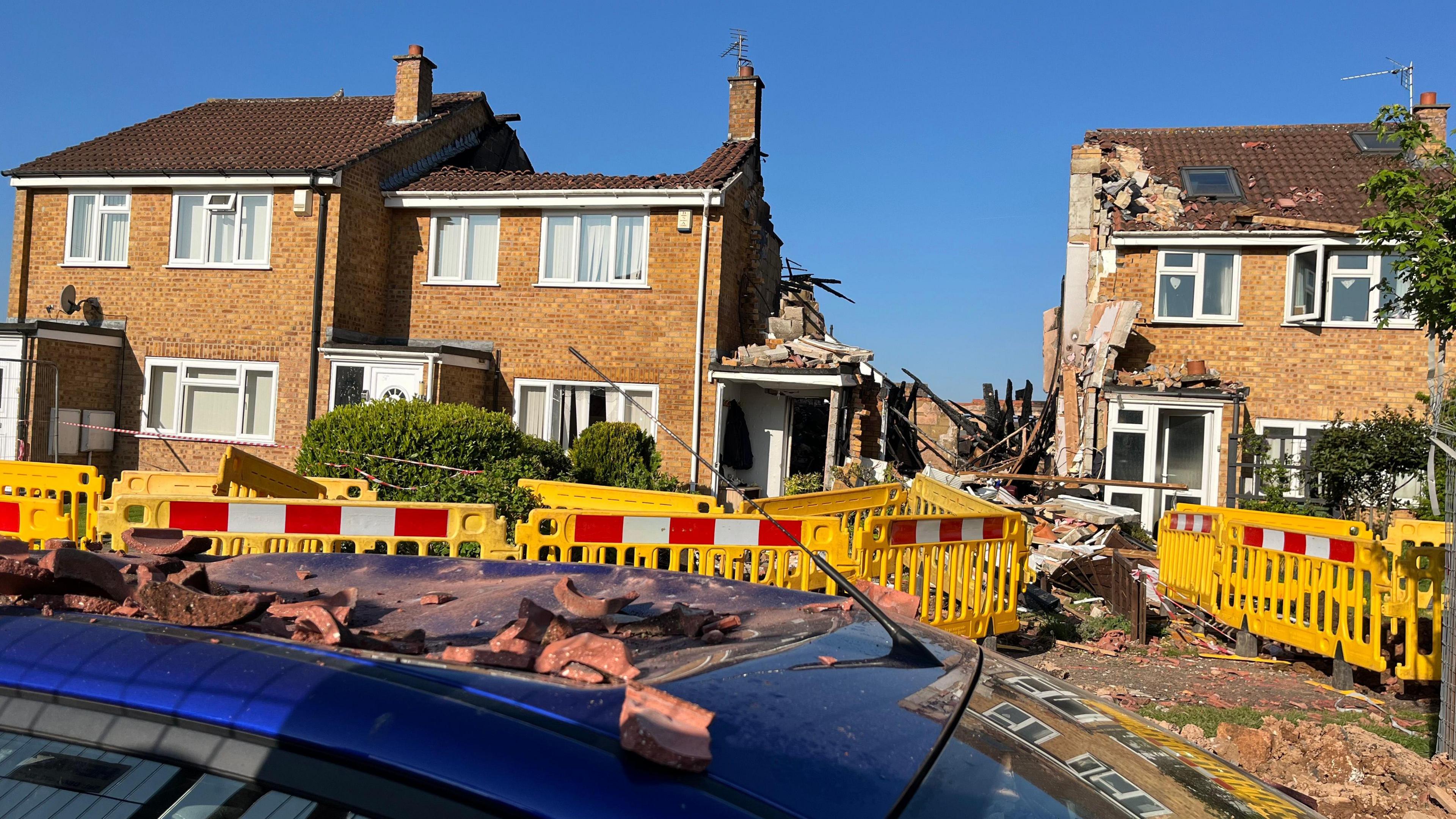 A row of four houses, one of which has been burnt to the ground. The house to the right of it has had it's roof taken off. There is a yellow cordon surrounding the houses. In front of the house is a car that has been destroyed by rubble.