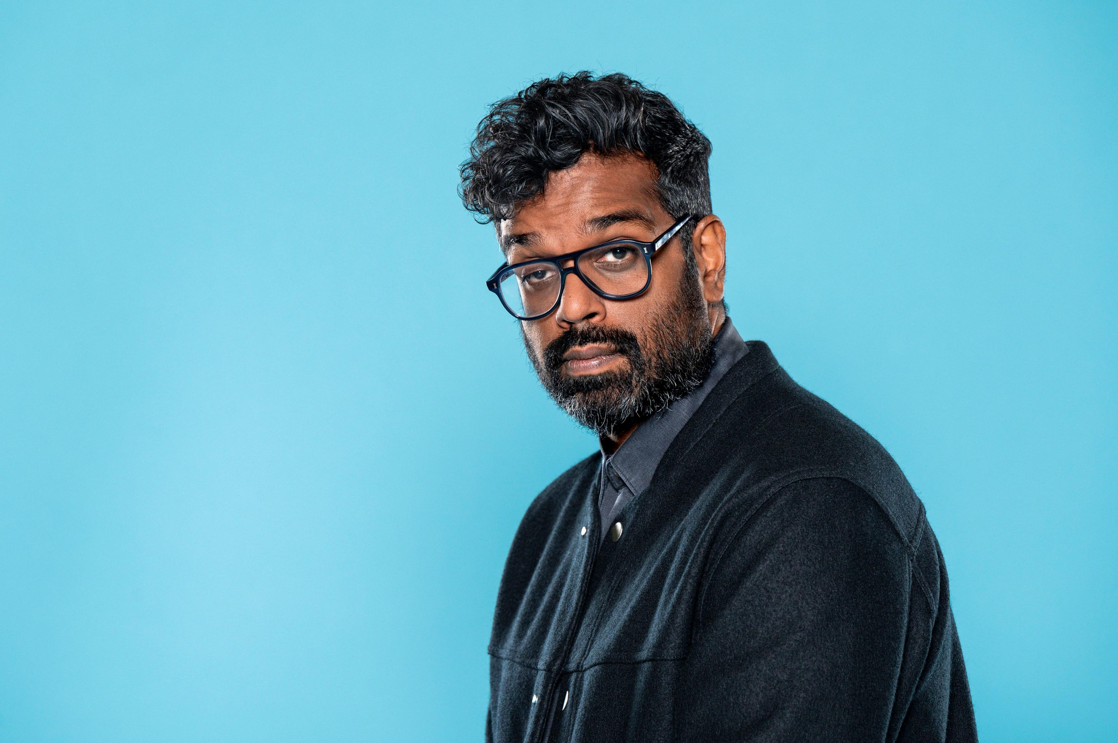 A man, Romesh Ranganathan, in a suit, dark shirt and glasses