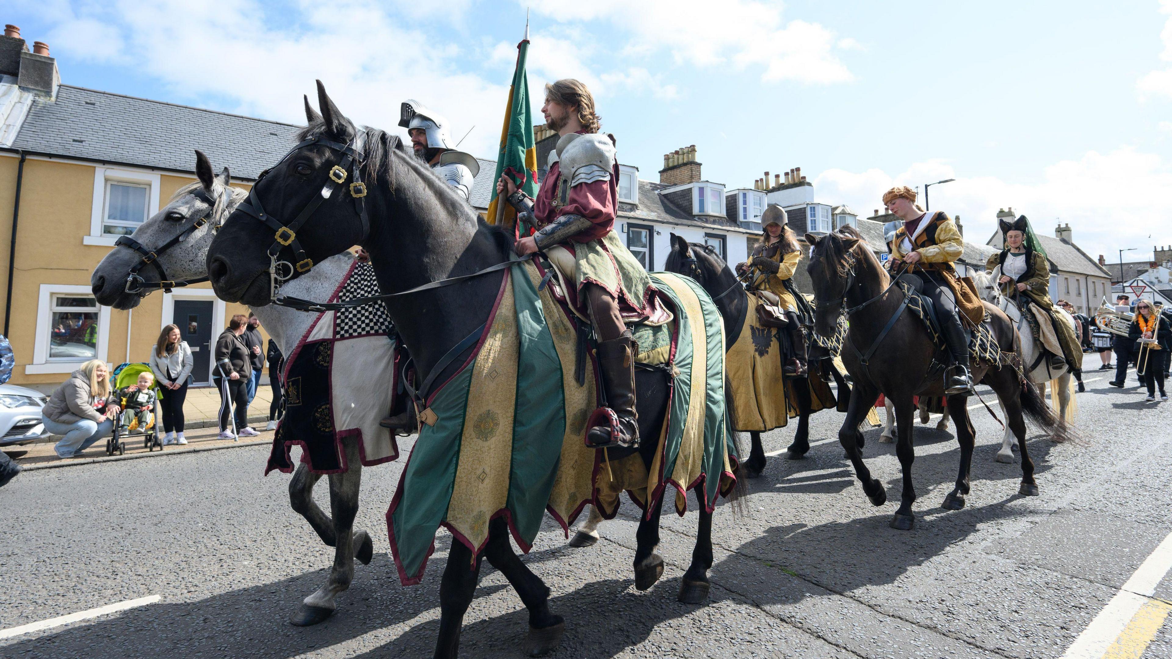 Several men and women on horses, dressed in medieval clothing like knights and maids, riding along a street as part of a parade.