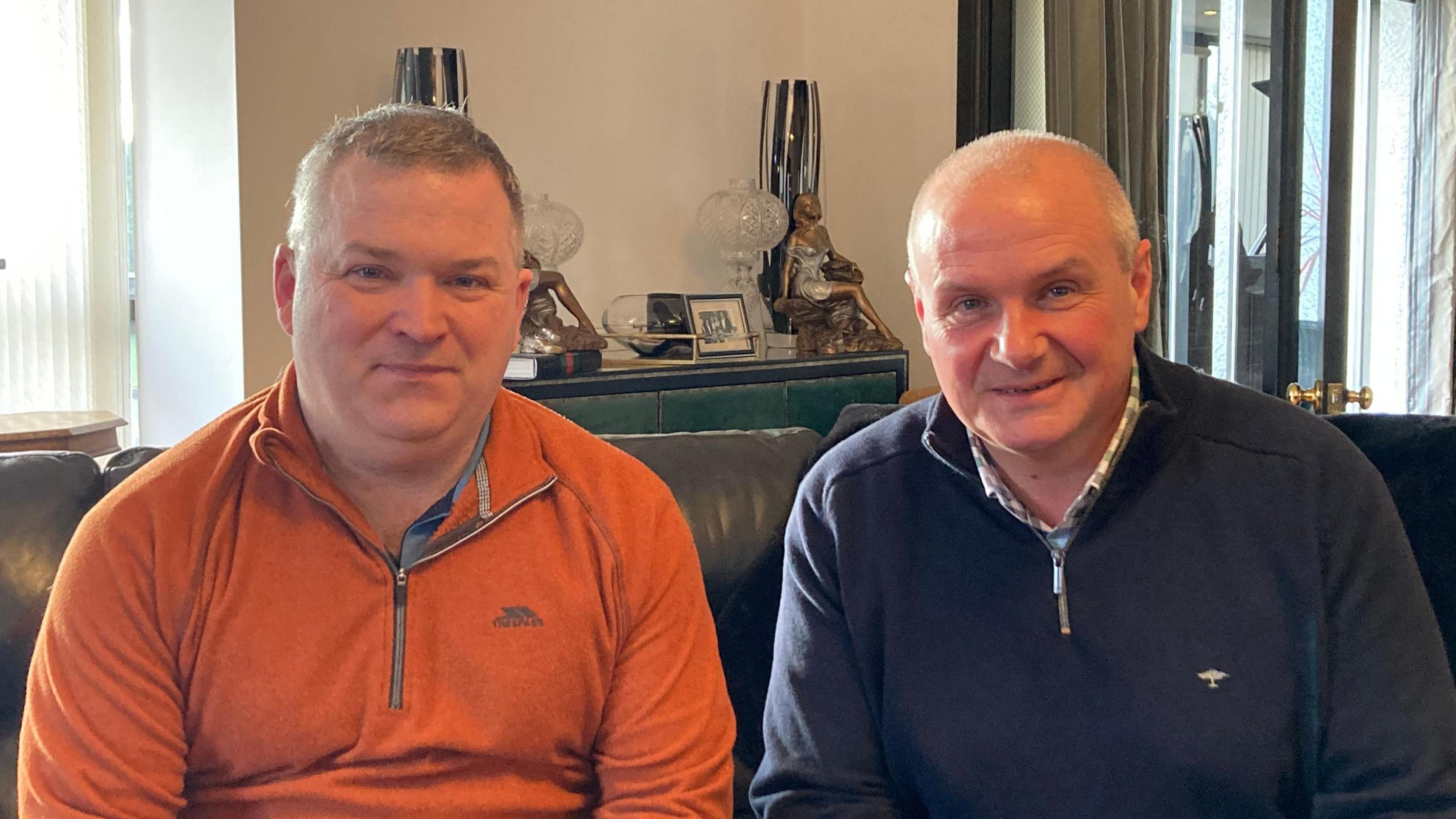 Paul McCaughey on the left, is wearing an orange sweater and looking at the camera. He has grey hair. Paul McAdam is on the right, wearing a dark jumper with a check shirt underneath. Both are sitting down and looking into the camera.