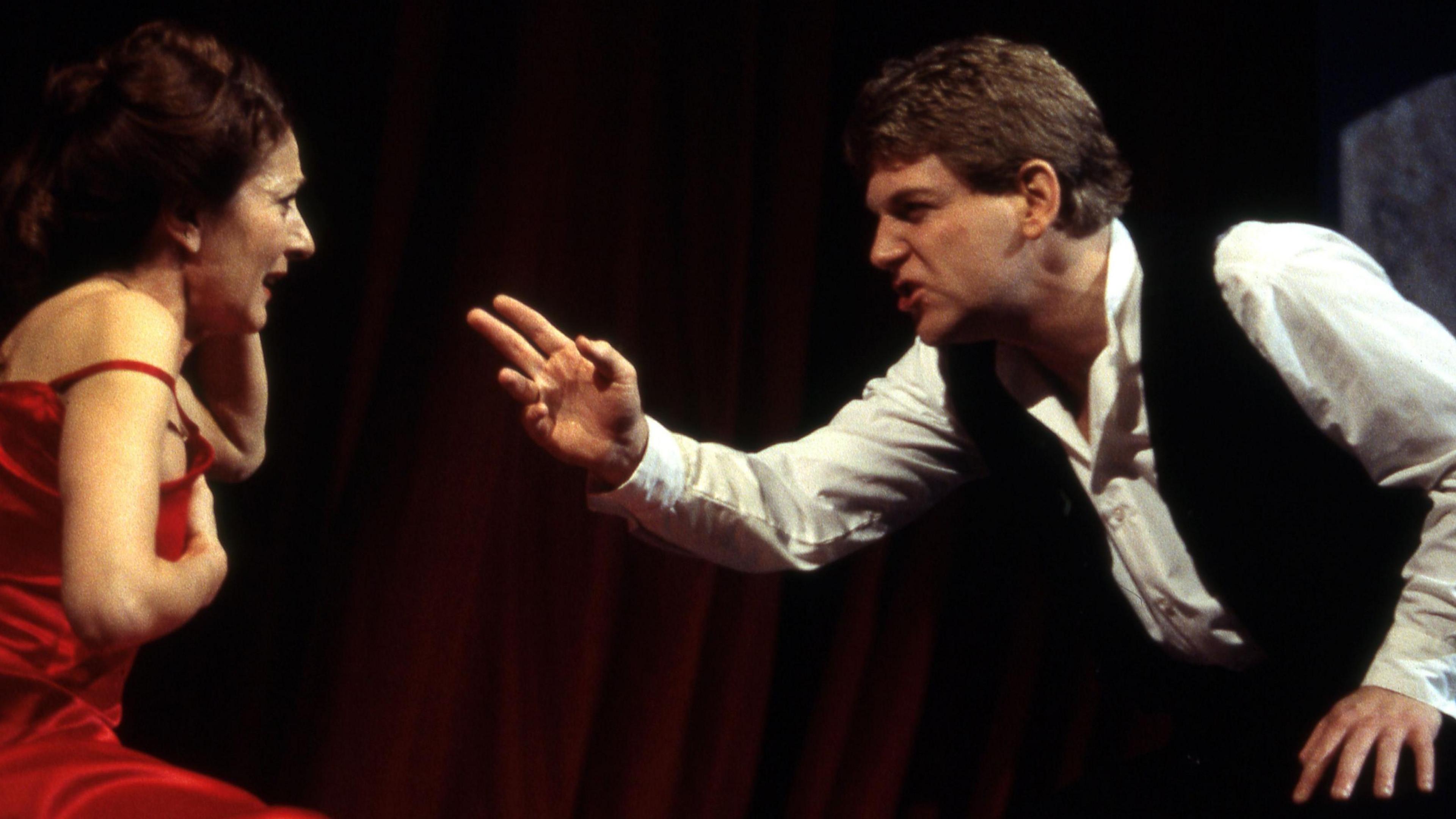 Side profile of Jane Lapotaire with brown hair in a bun (playing Getrude) in red sleeveless dress with strap coming off her shoulder with an anguished expression being berated by Kenneth Branagh's Hamlet, dressed in white shirt and dark waistcoat -- who's gesticulating with one arm at Gertrude. 