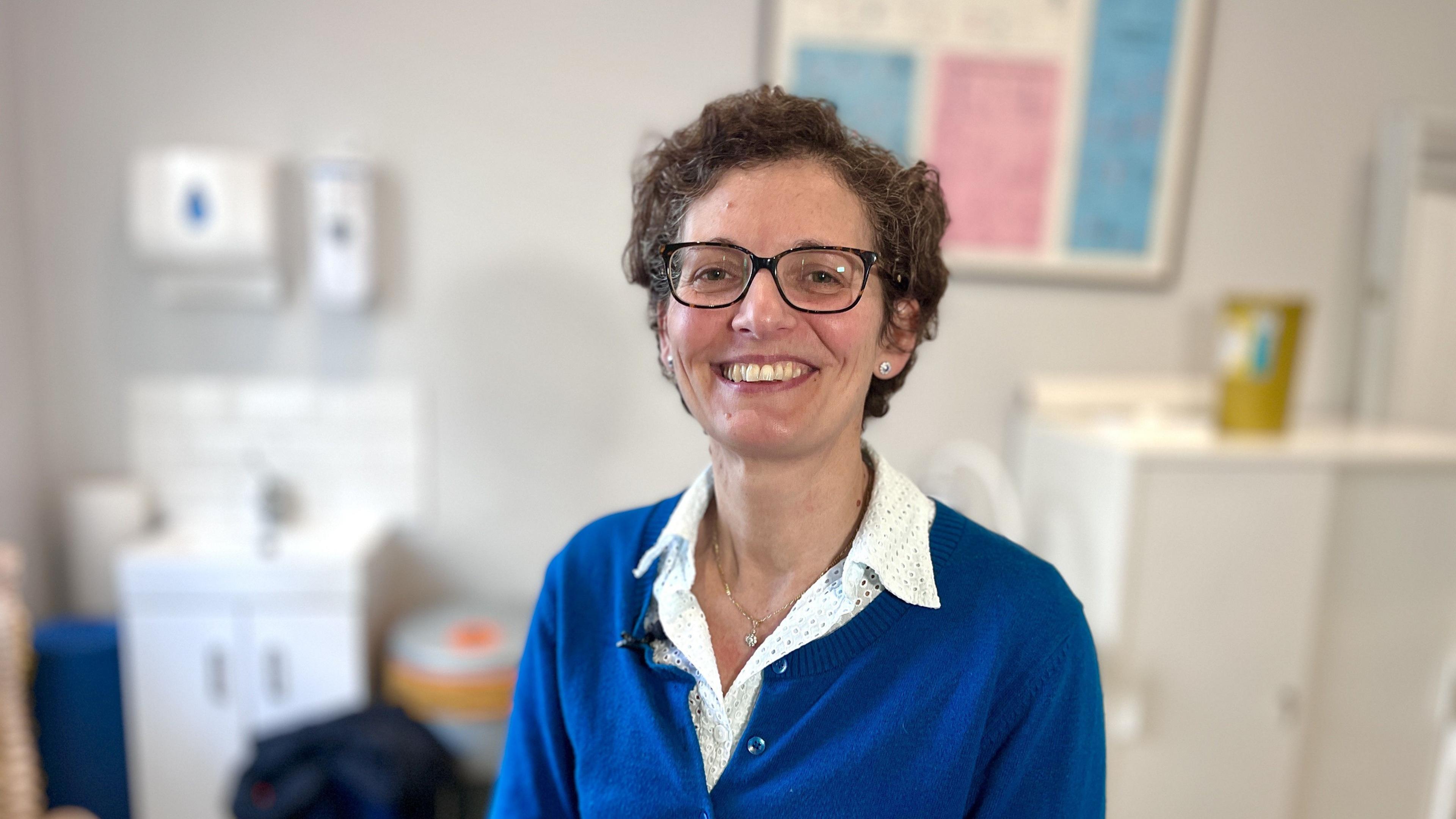 Dr Yvonne Girgis-Hanna looks directly at the camera and smiles. She is wearing tortoiseshell glasses and a blue cardigan over a white patterned blouse and is sitting in a consulting room.