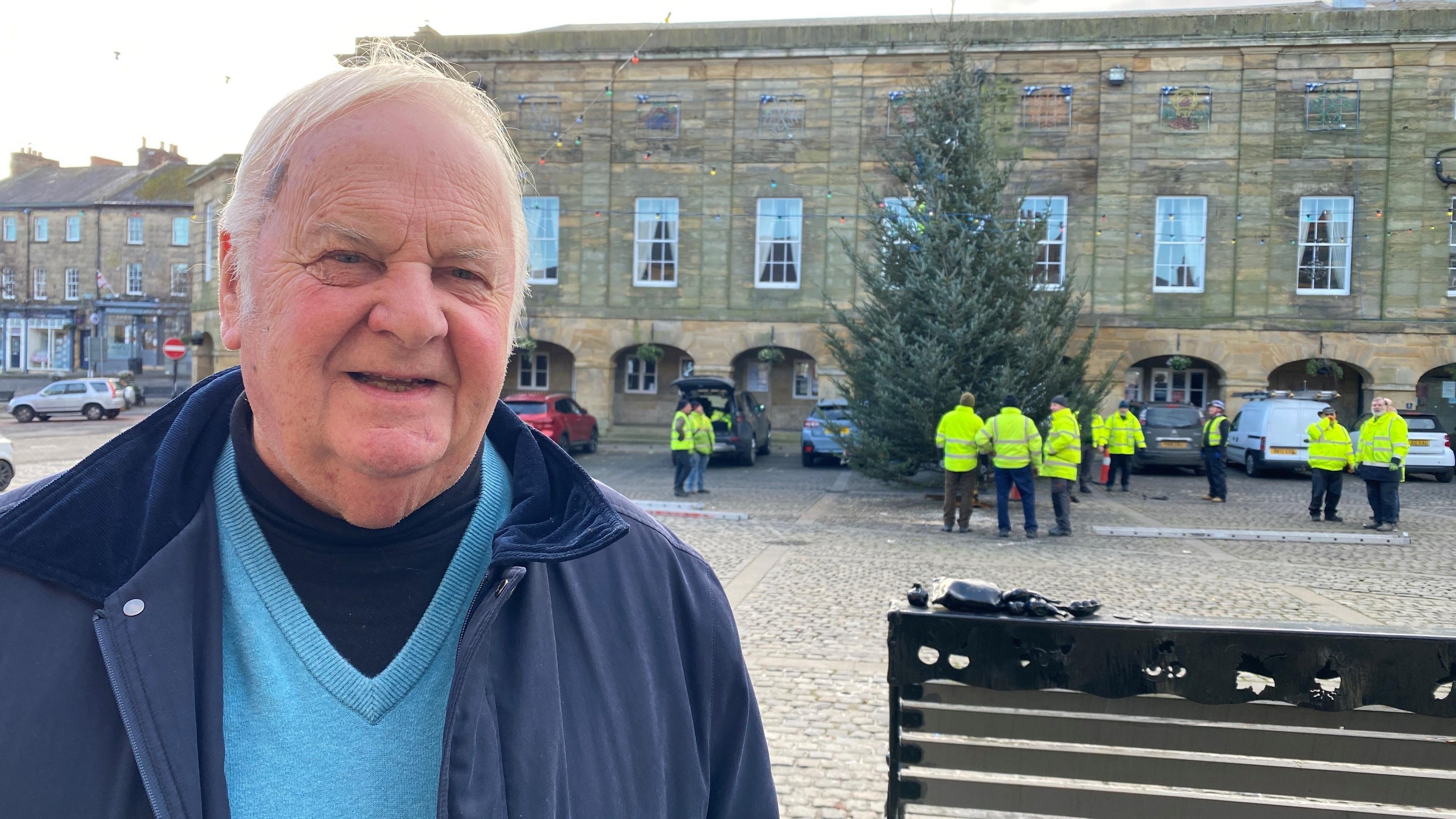 An older man wearing a blue coat and light blue jumper is standing in front of the Christmas tree 