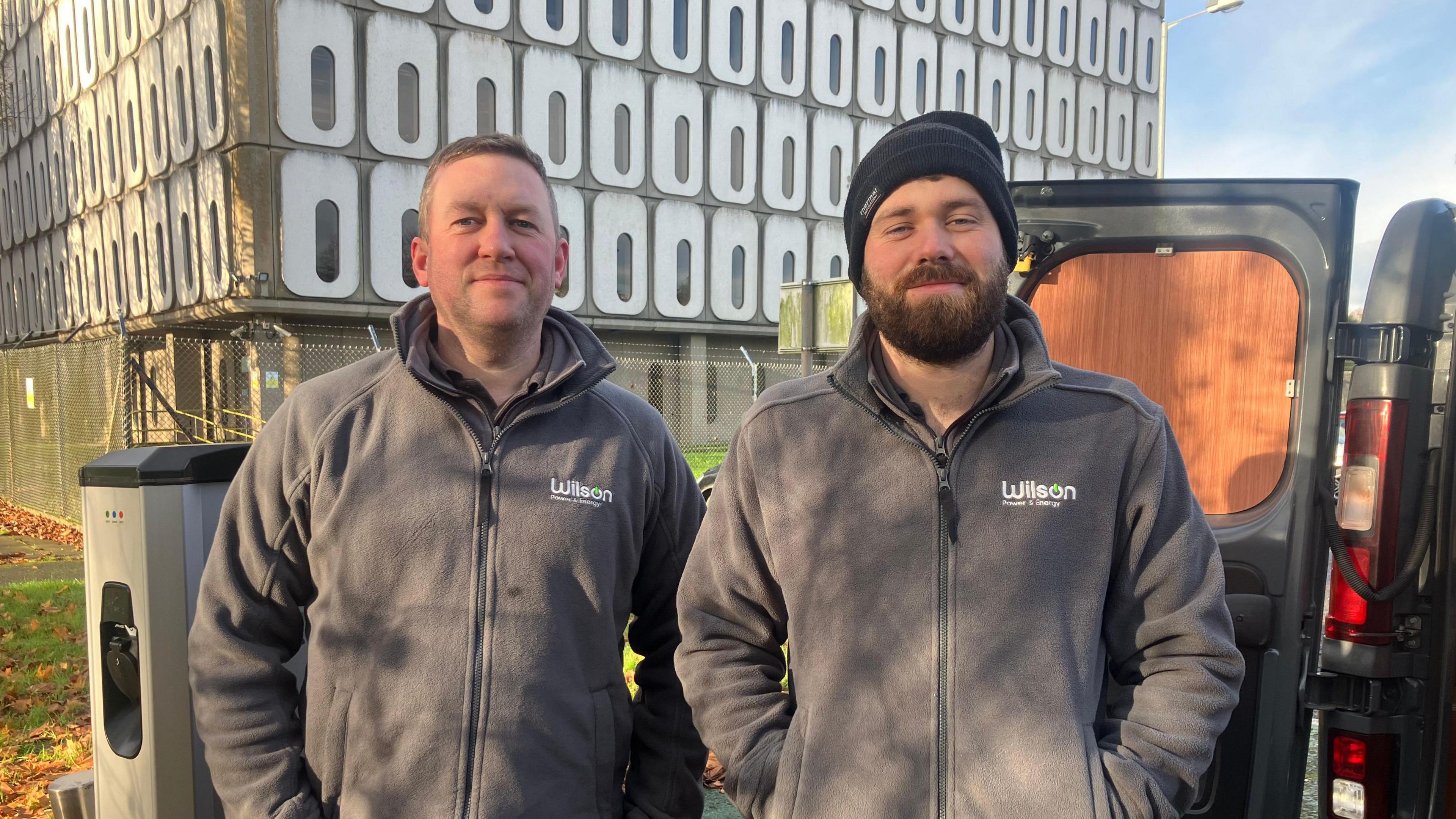 David and Stephen stand in front of Marlborough House. They are both wearing hoodies saying Wilson abd one is wearing a hat. The door to the back of their van is open.