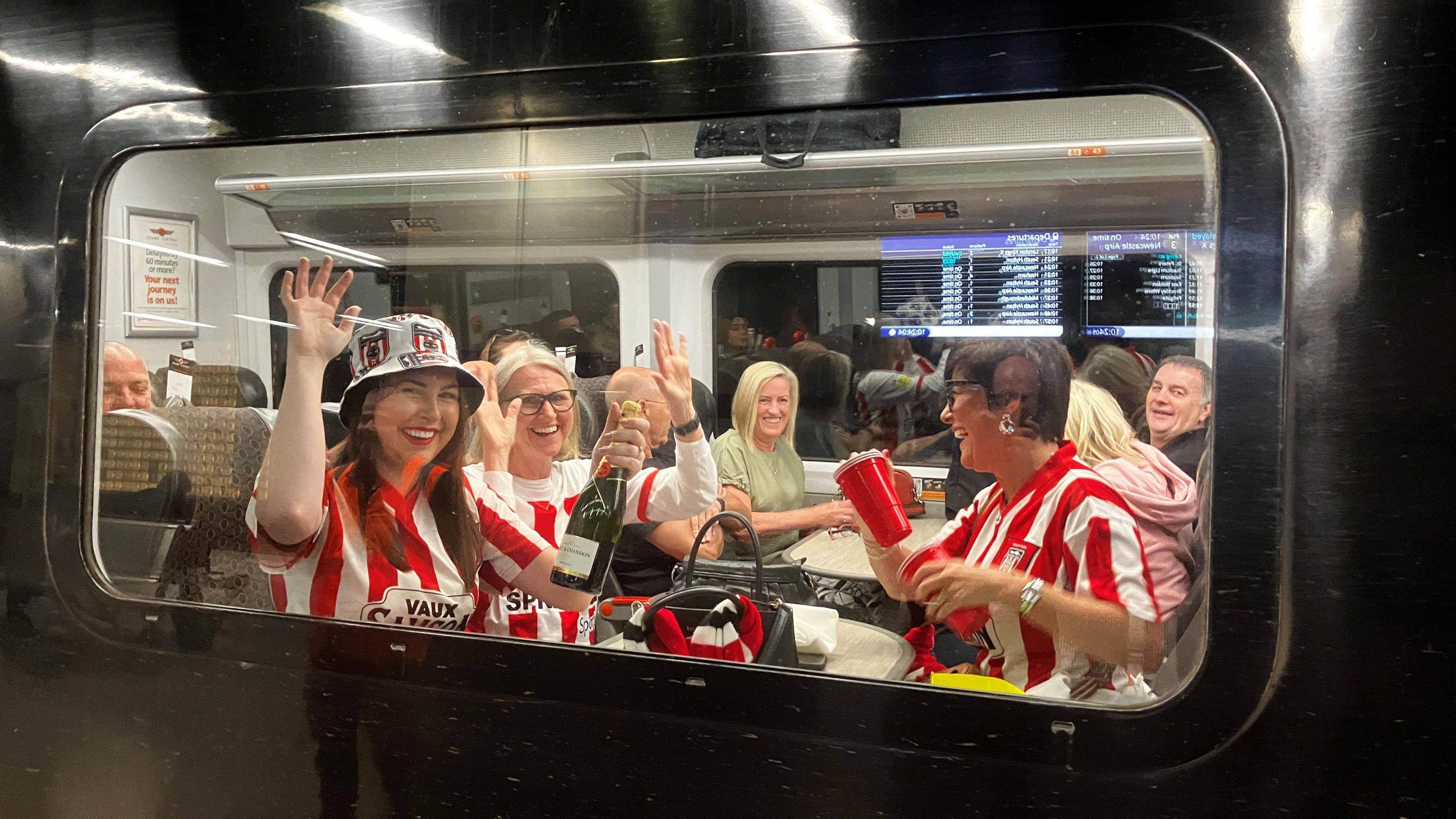 A group of women wearing red and white Sunderland shirts and holding a bottle of bubbly inside a train carriage. Everyone is smiling.
