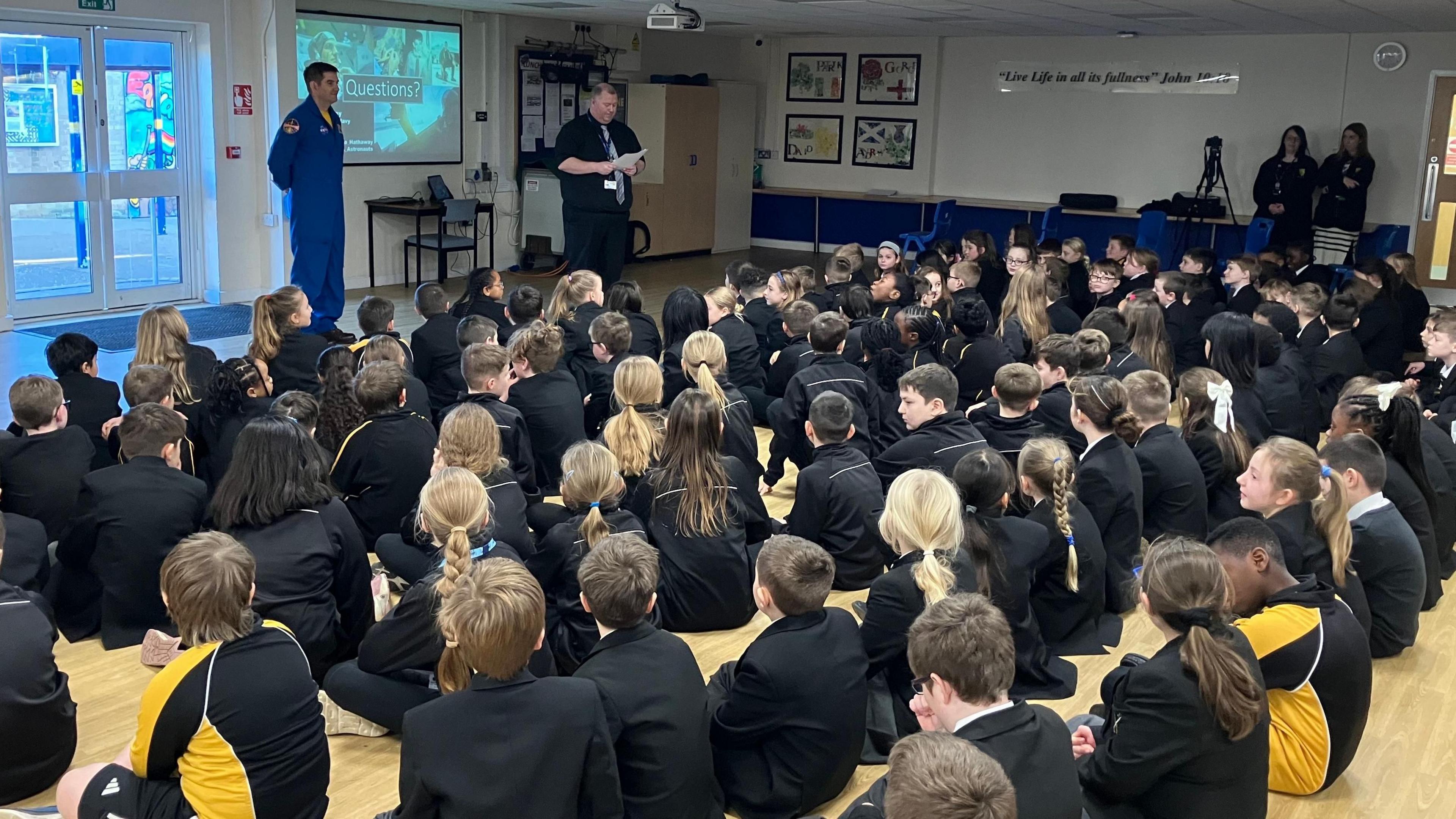 Commander Jack Hathaway is standing at the front of a school hall wearing blue NASA overalls with some doors behind him and a projector screen to the side of him. The screen has the word "Questions?" on it. In front of him are dozens of school children sitting on the floor looking at him, with their backs to the camera.