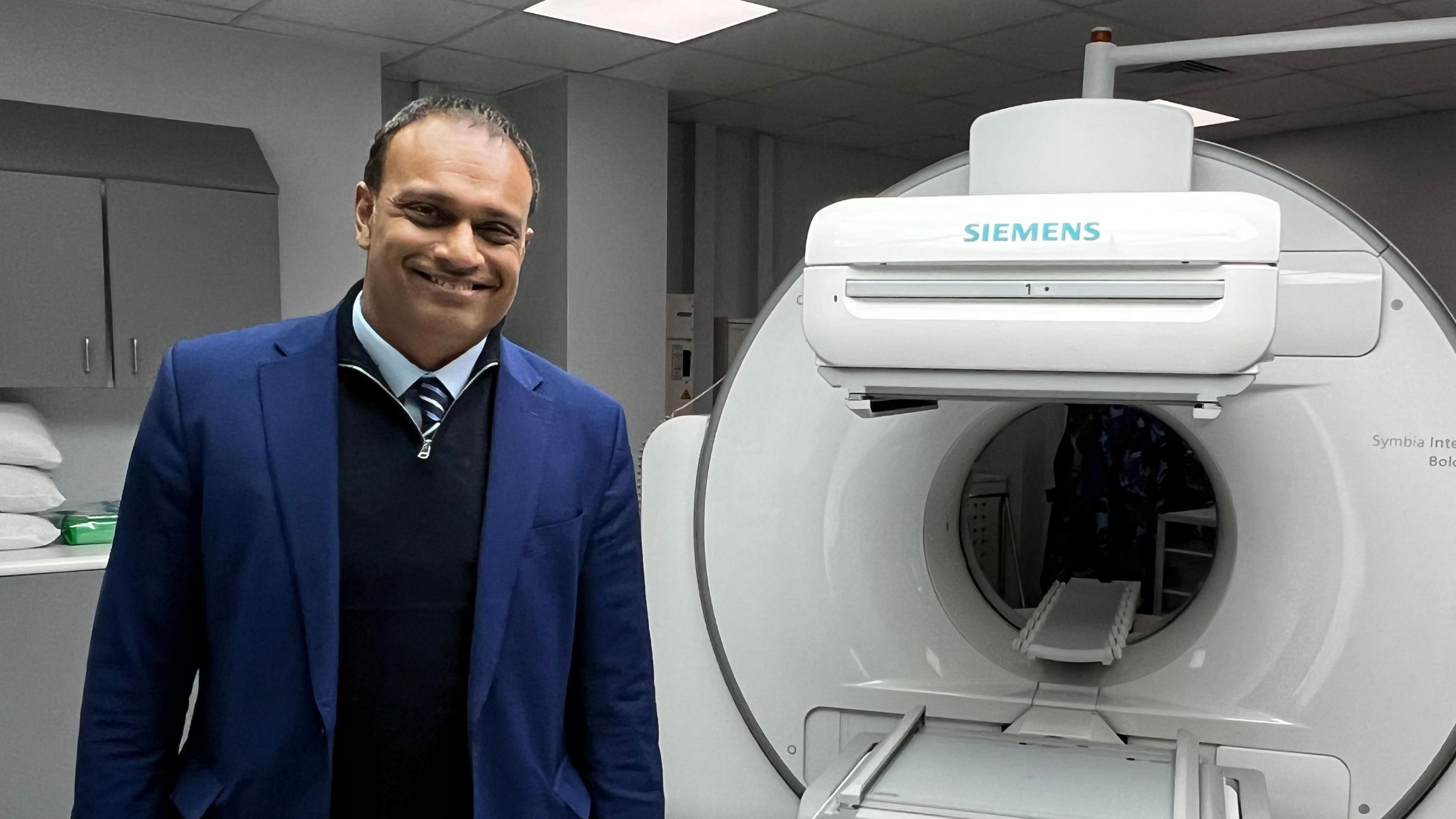 Dr Randeep Kulshrestha stands in front of a medical scanner. He wears a blue suit, with a blue shirt, striped tie and navy quarter zip jumper. Behind him is a large circular white scanner. 