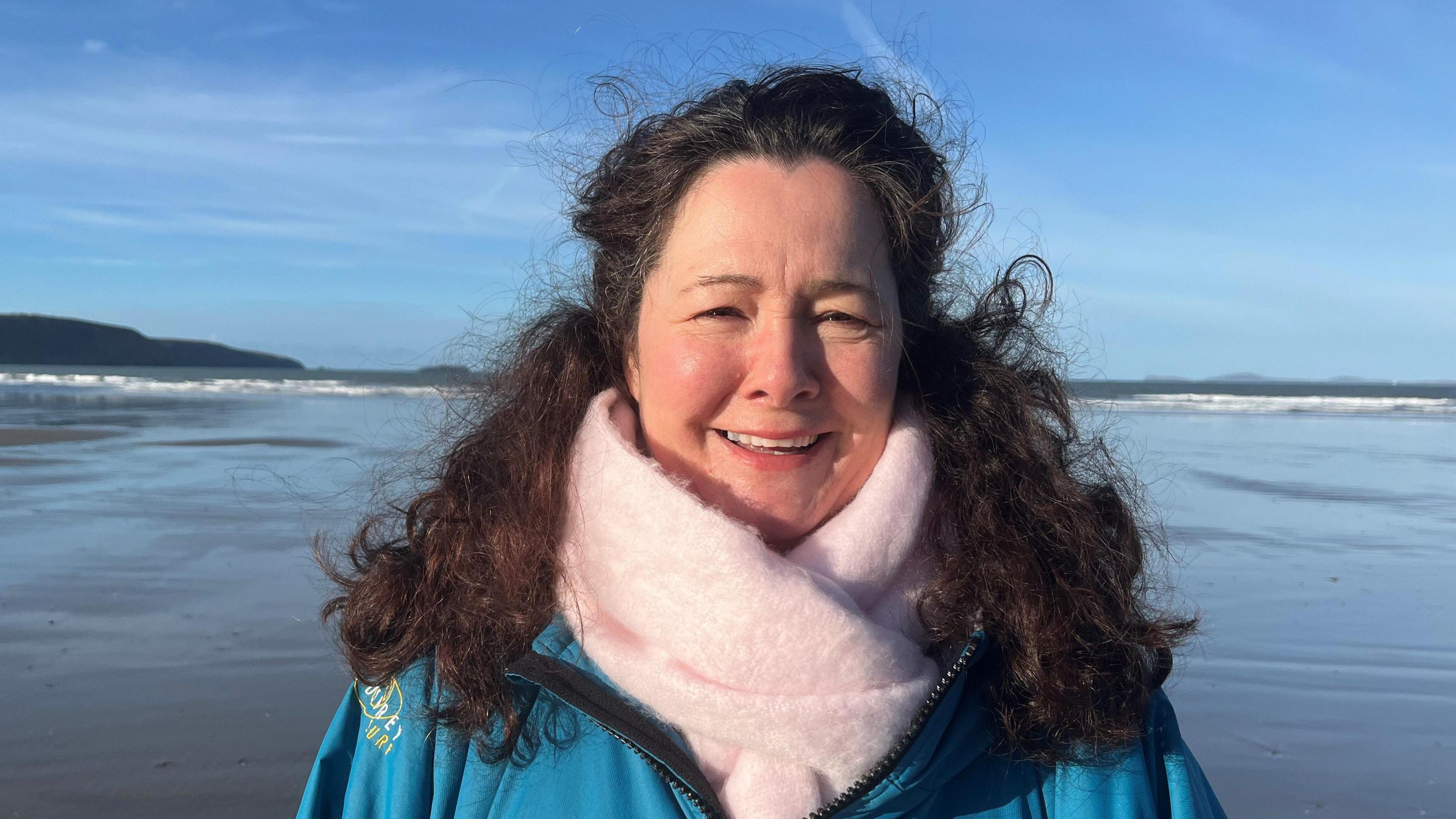 Kate Evans smiles at the camera. She is stood on the beach with the sea and wet sand behind her, wearing a blue jacket and big pink scarf. 