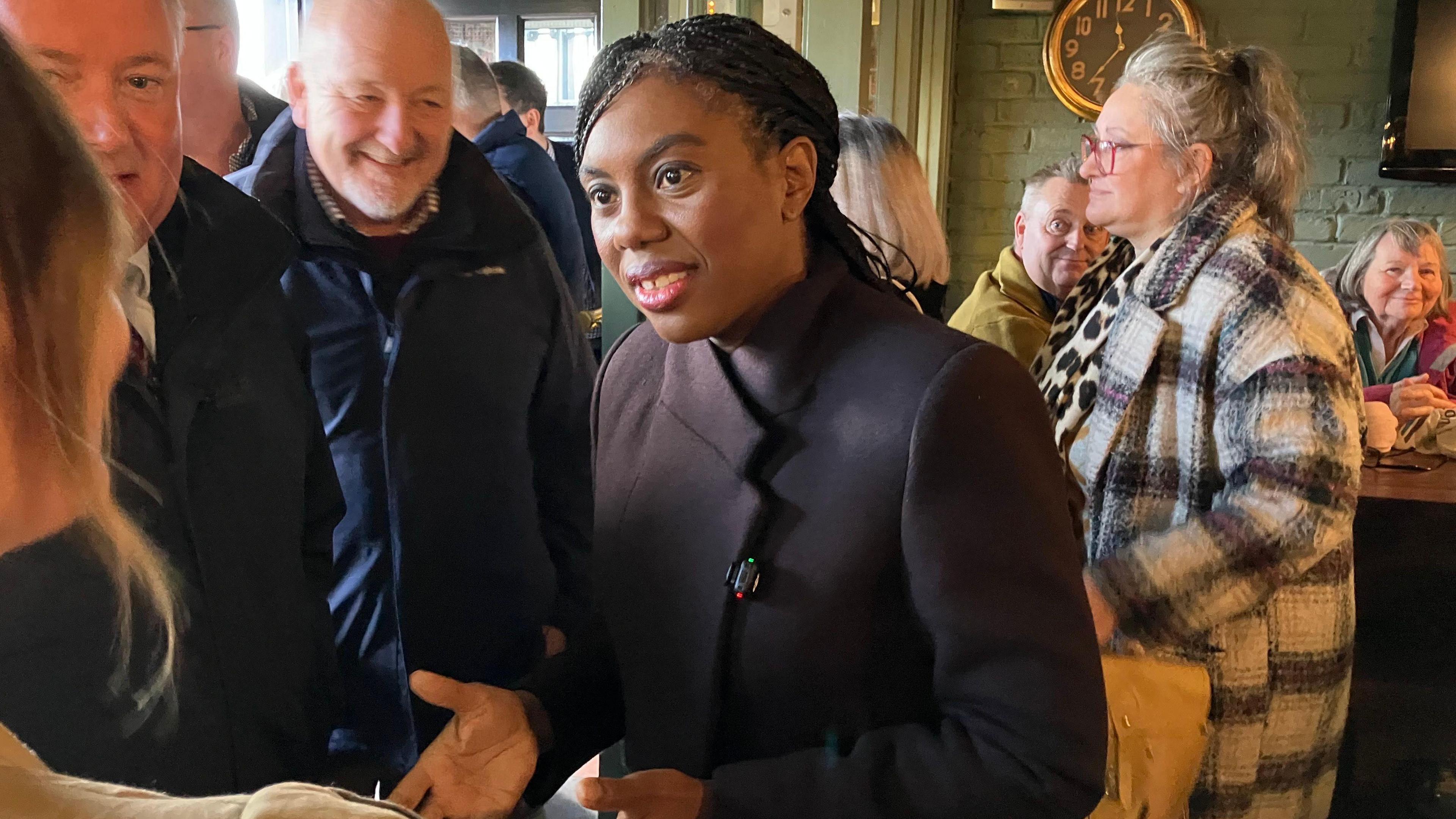 Kemi Badenoch speaking to a business owner in a busy bar restaurant in Monmouth. On her right hand side are local Senedd member Peter Fox and Conservative Senedd group leader Darren Millar. All three are smiling and wearing dark coats.