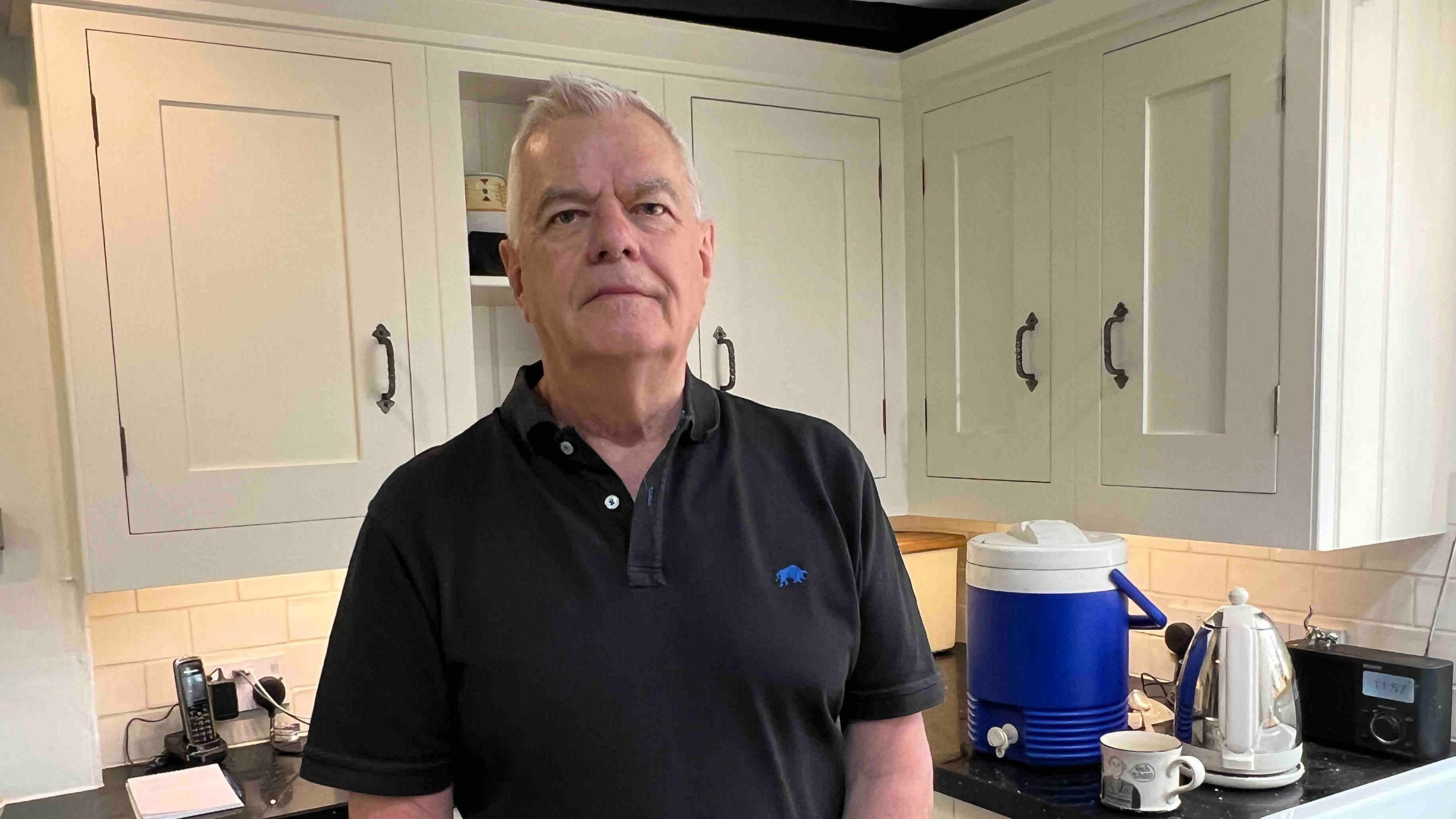 A man in a black polo top standing in a kitchen with white cupboard doors and a black granite work surface.