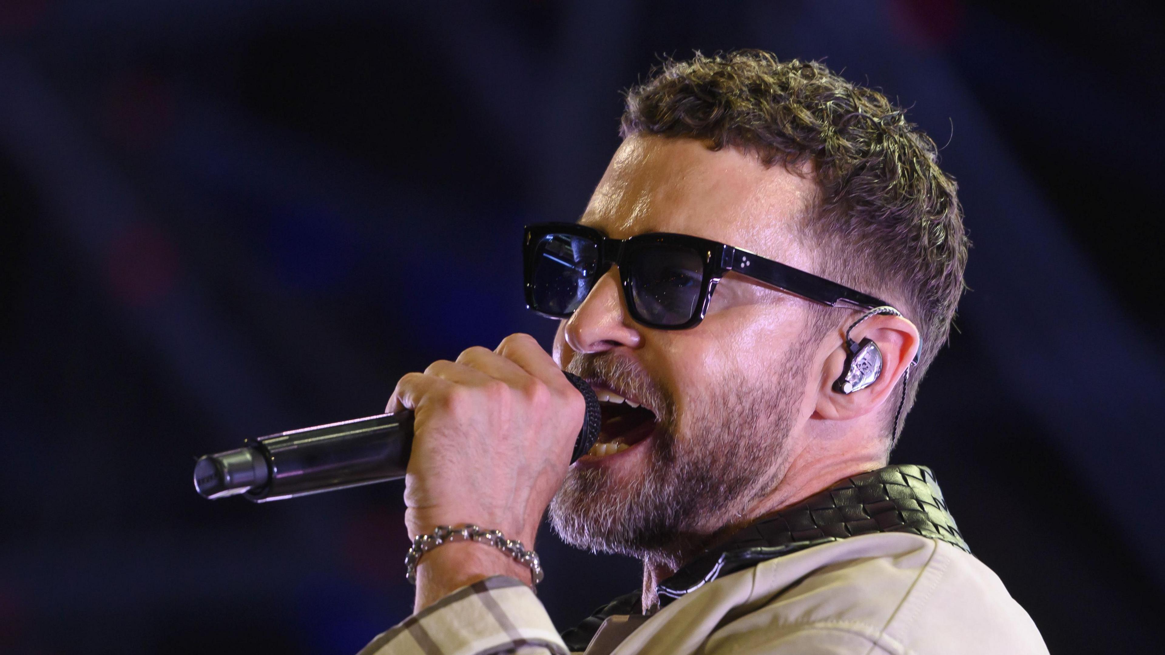 Justin Timberlake is wearing a pair of large sunglasses. He holds a microphone up to his mouth and is singing into it. His hair is brown, curly and cut short. He has a beard. 