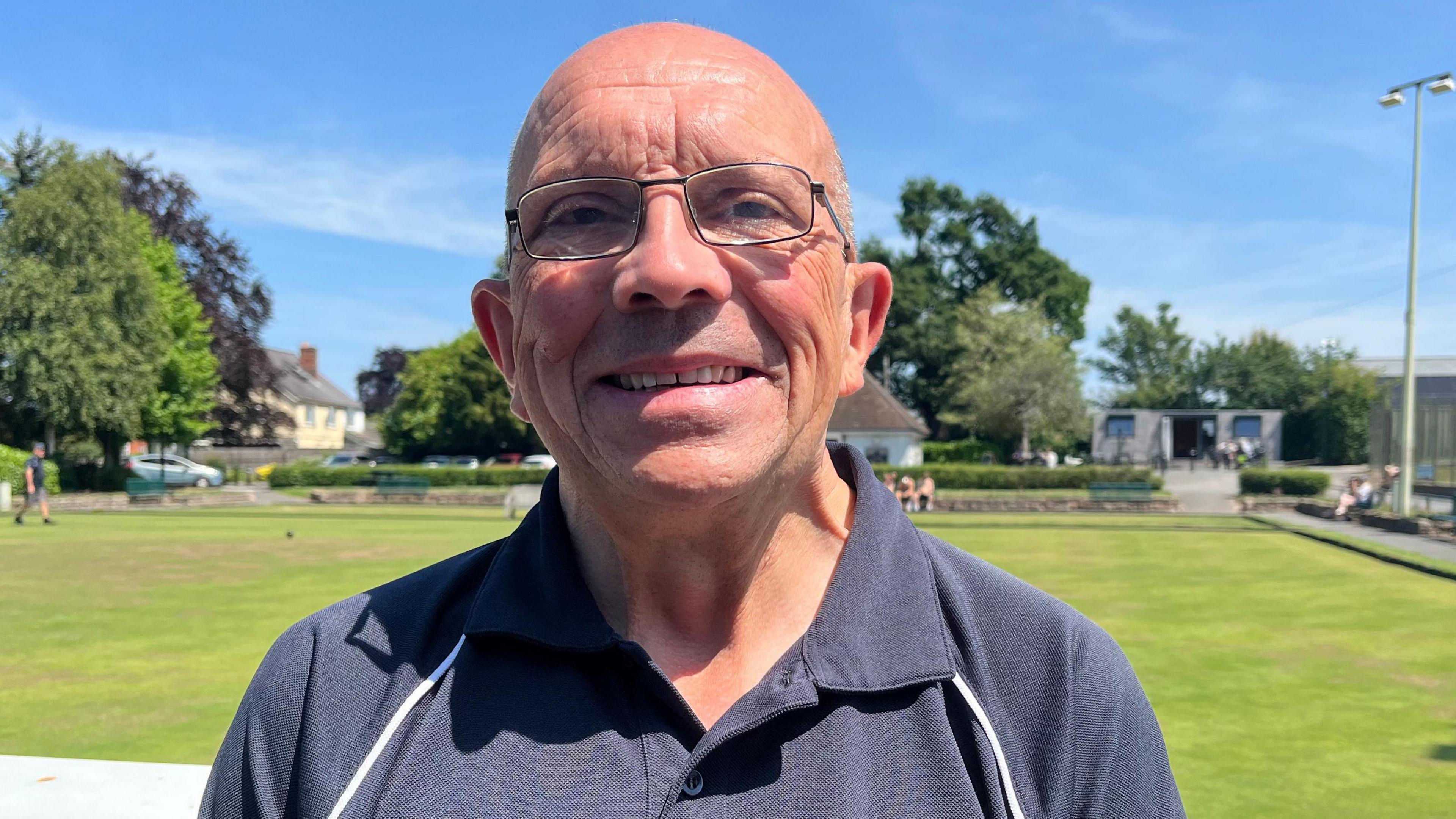 Mark looking into the camera and smiling. He has a bald head and is wearing a navy blue short-sleeved sports shirt. He is standing in front of a bowling green, which is green and grassy. The sky is bright blue with a few clouds, and it is very sunny.