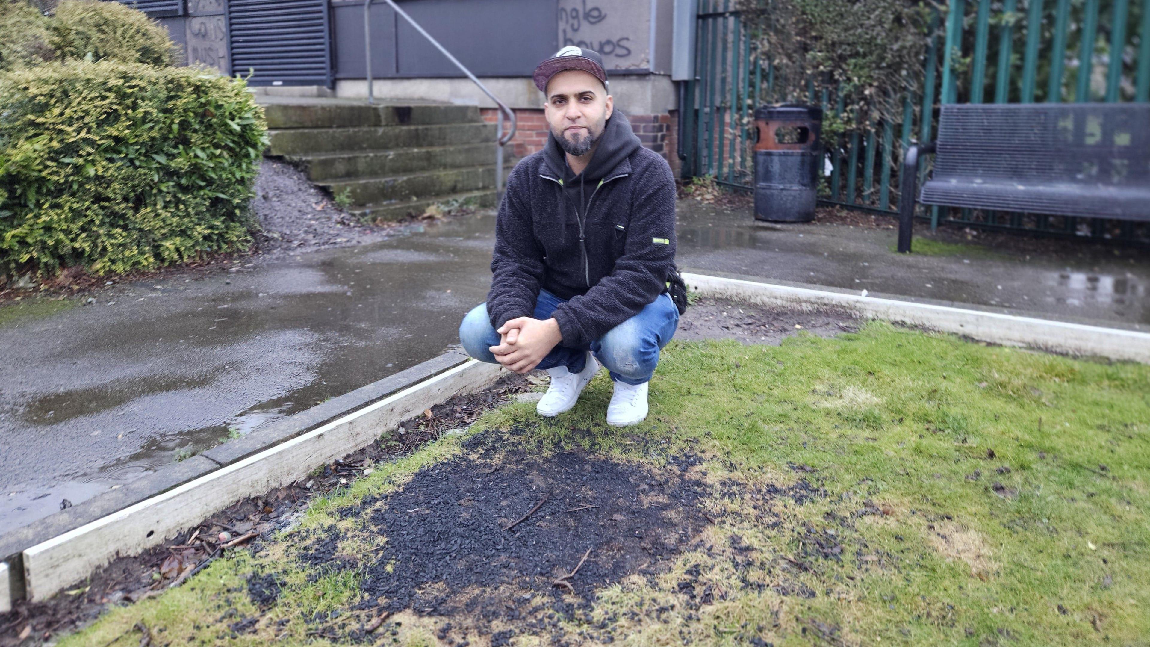 A man crouches by a piece of blackened grass in a park. Behind him is a park bench and a building with some graffiti scrawled on it.