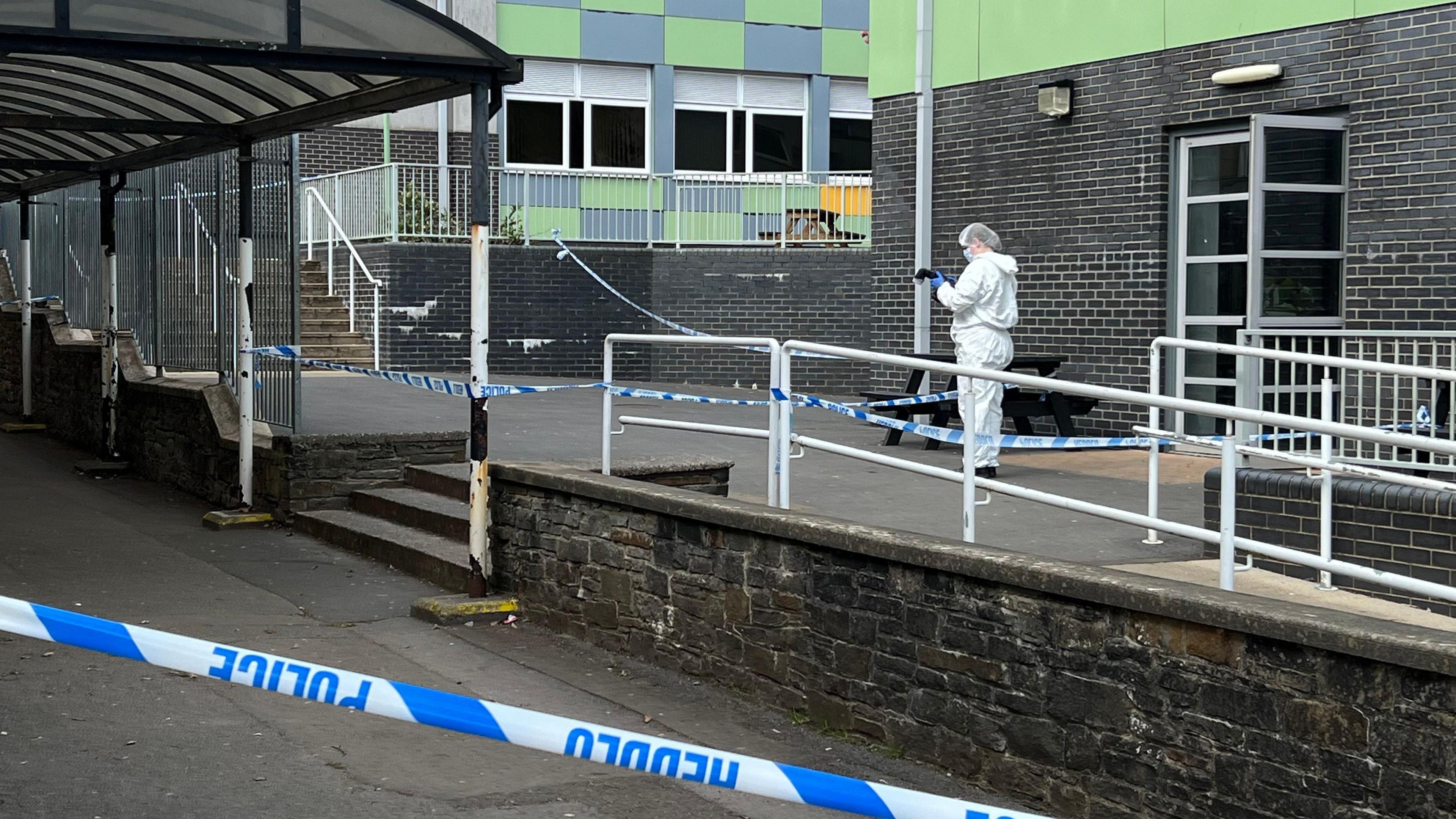 Police tape on a landing outside the school where a forensic officer in all white jumps suit, hair net and mask with a long lens camera