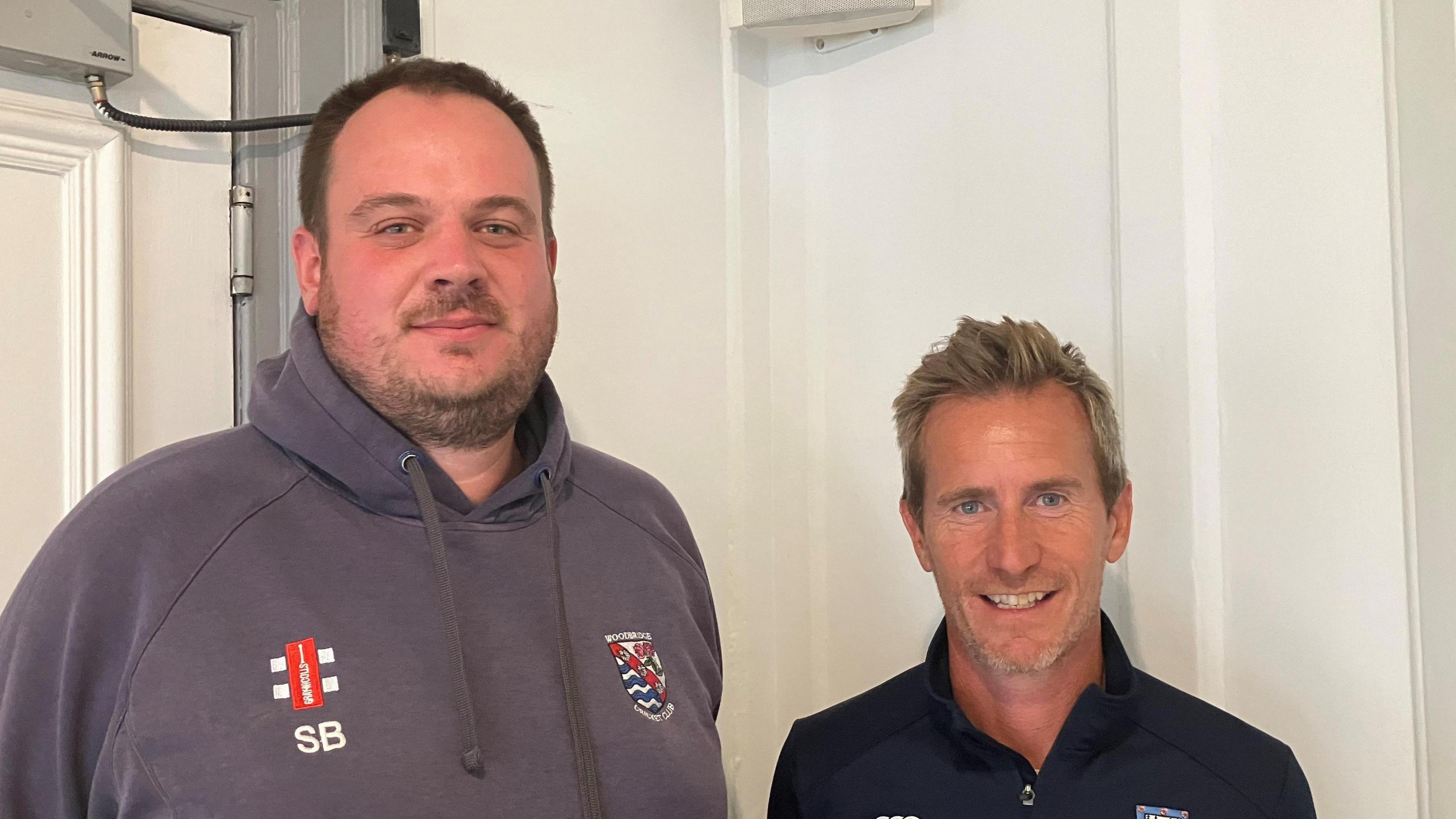 Scott Button stands next to Ralph Pruden. Scott has short dark hair and wears a dark grey hoodie, with his initials on one side of his chest and the cricket club's logo on the other side. Ralph has short light hair and wears a navy sweater. 
