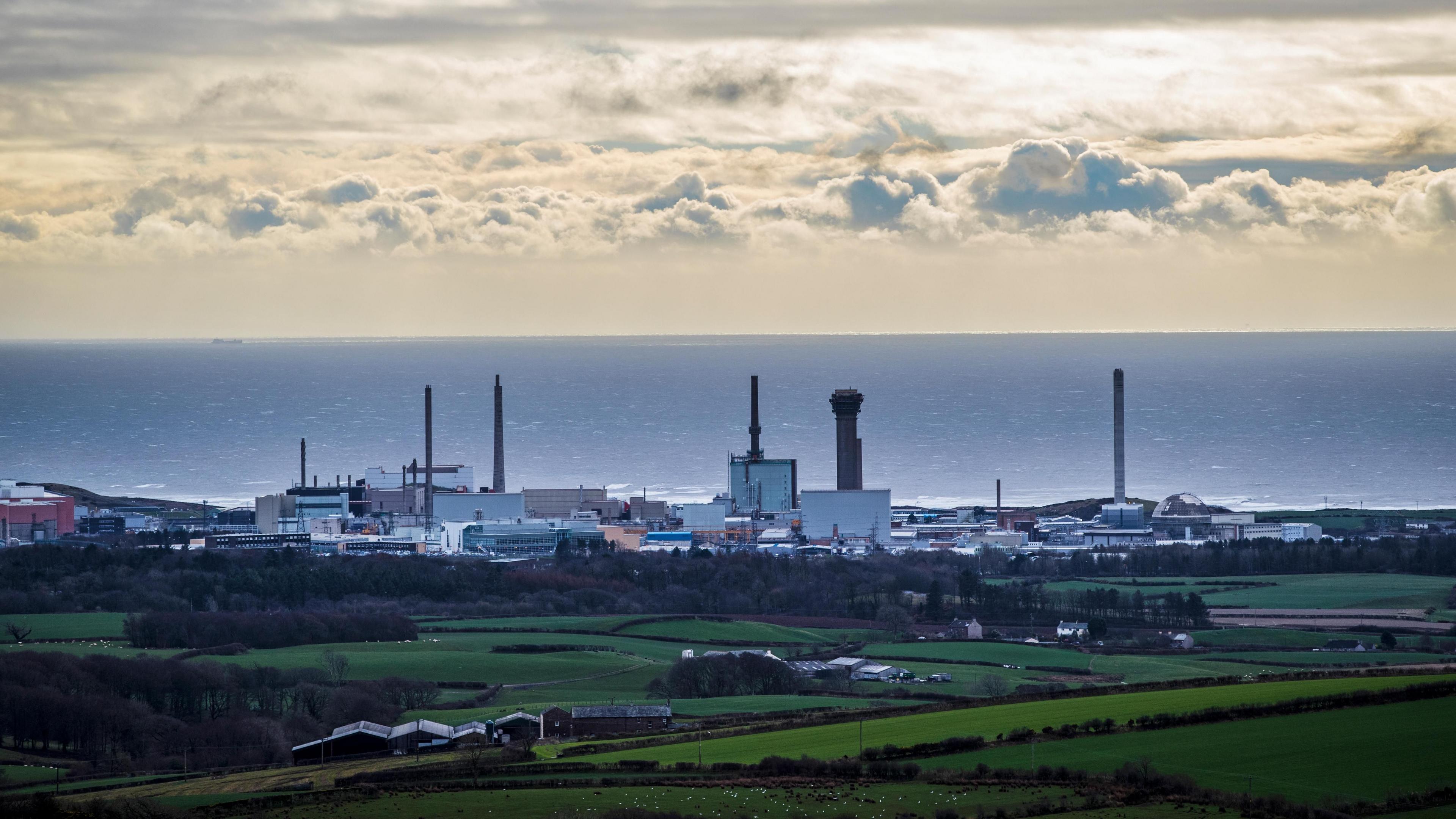 A view of Sellafield nuclear site in West Cumbria from a distance, looking out towards the coast. fields are visible in the foreground with the sea in the background beyond Sellafield, which is a complex of buildings of varying heights.