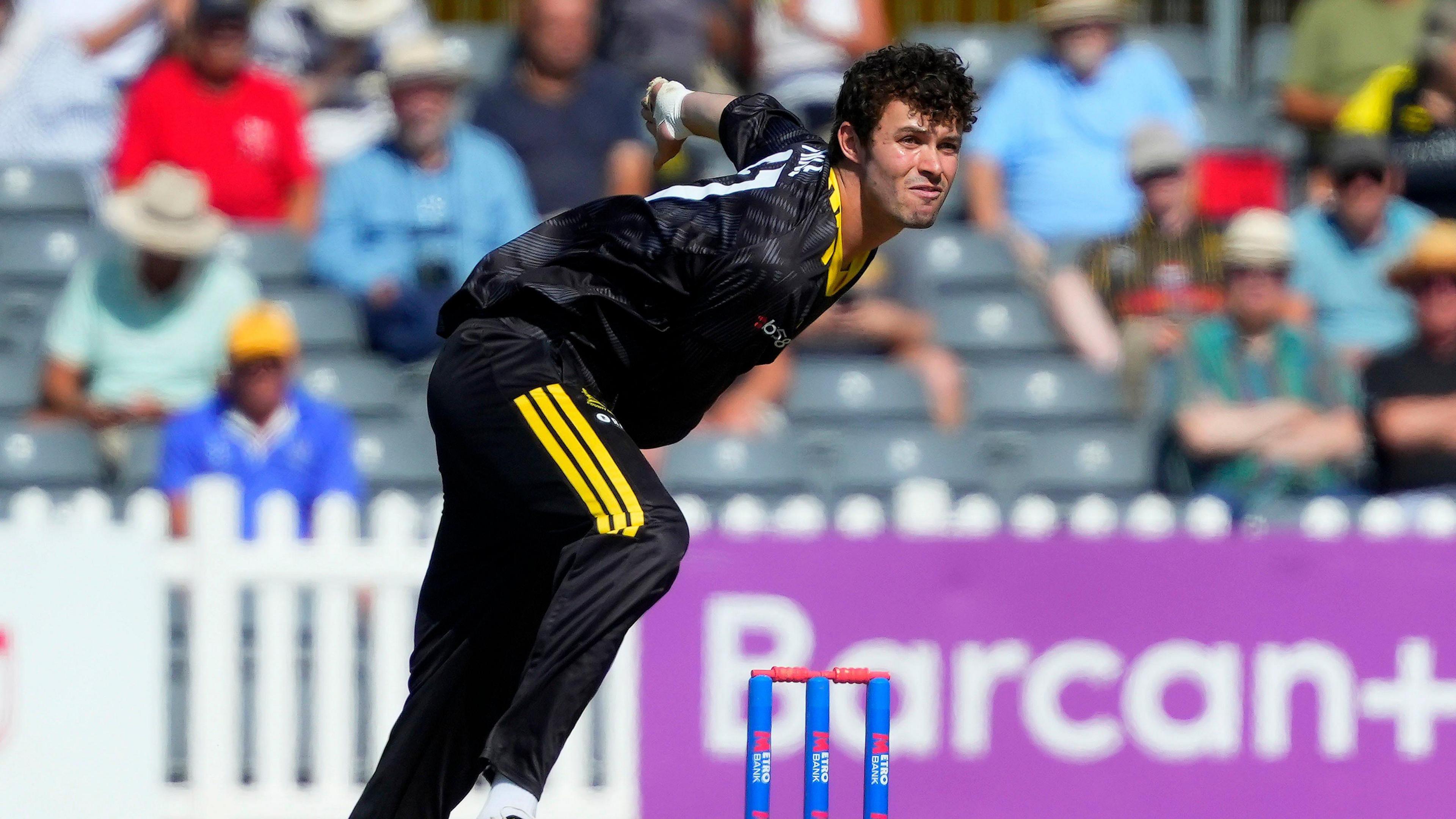 Ollie Price bends forwards after bowling a ball during a match for Gloucestershire. He is wearing black team kit