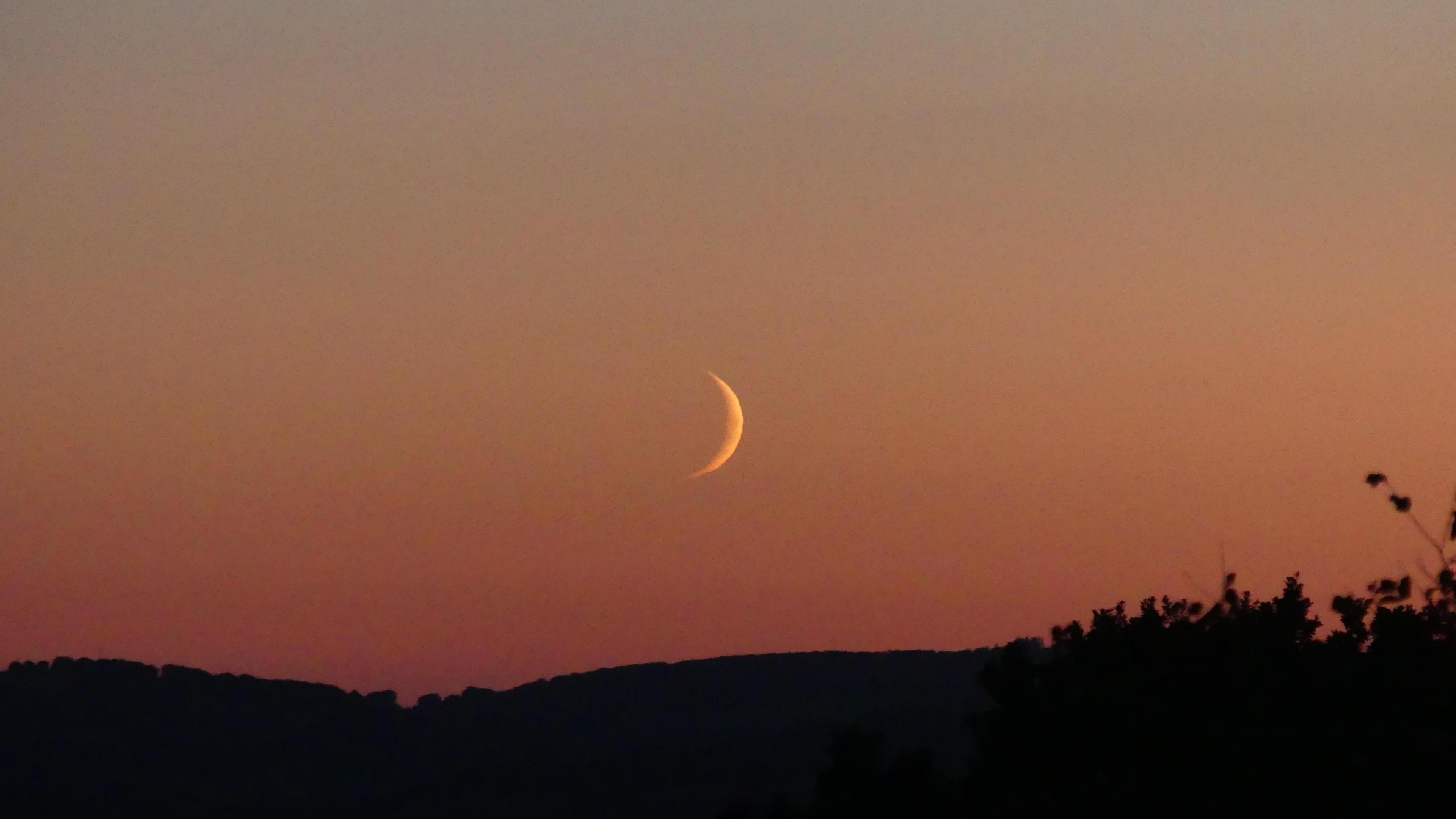 As the sun sets, a crescent moon rises over the Somerset hills. The sky is still lit in red, orange and yellow hues from the sun, although the sun itself is not visible.