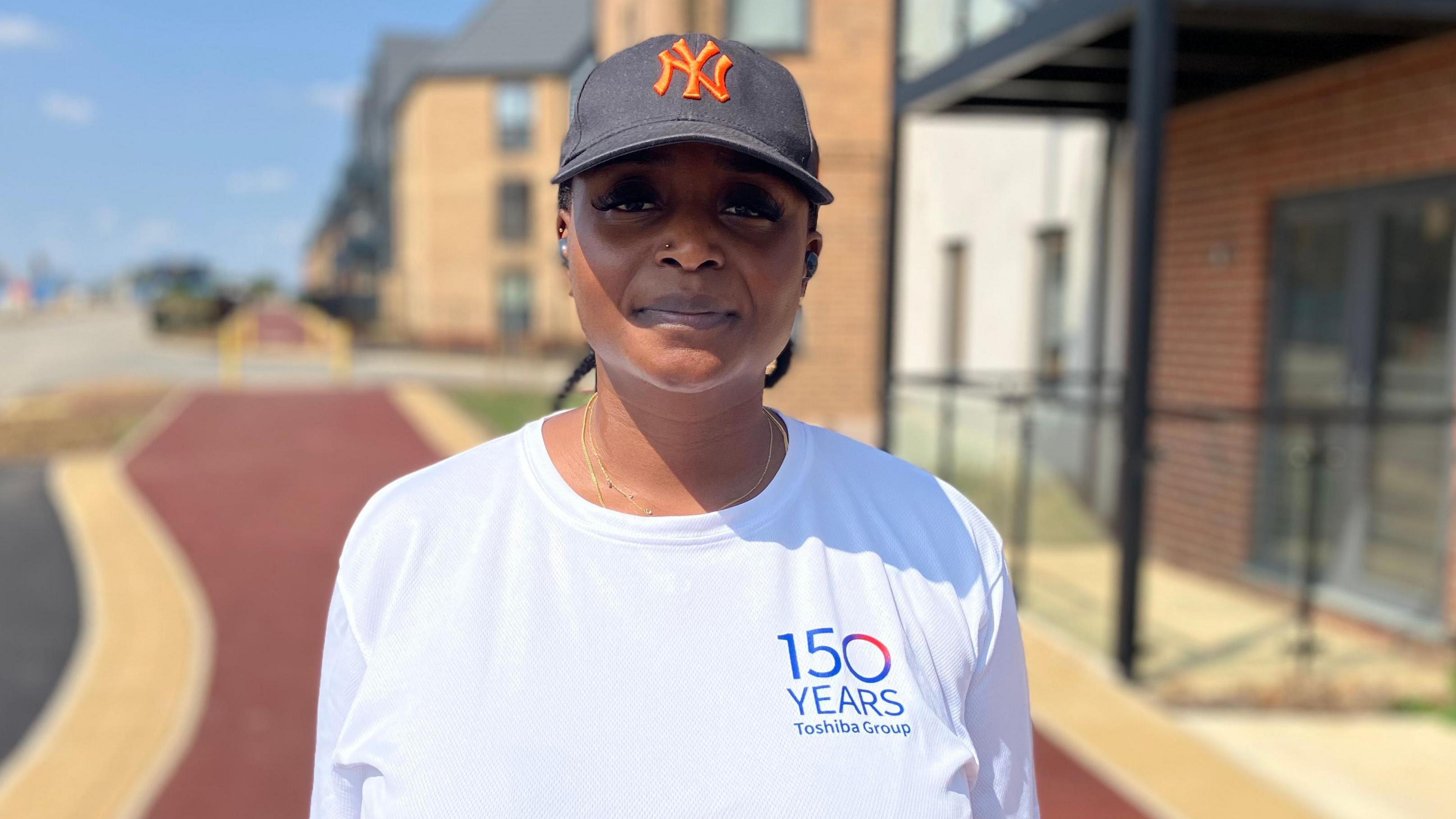 This is a head and shoulders picture of Mowale Jacobs who is looking at the camera and smiling. She is wearing a white T-shirt which says "150 years" on it. She is wearing a grey baseball cap with the orange letters "NY" on it. The background is blurred but shows a pathway on a modern residential streets with blocks of flats to the side.