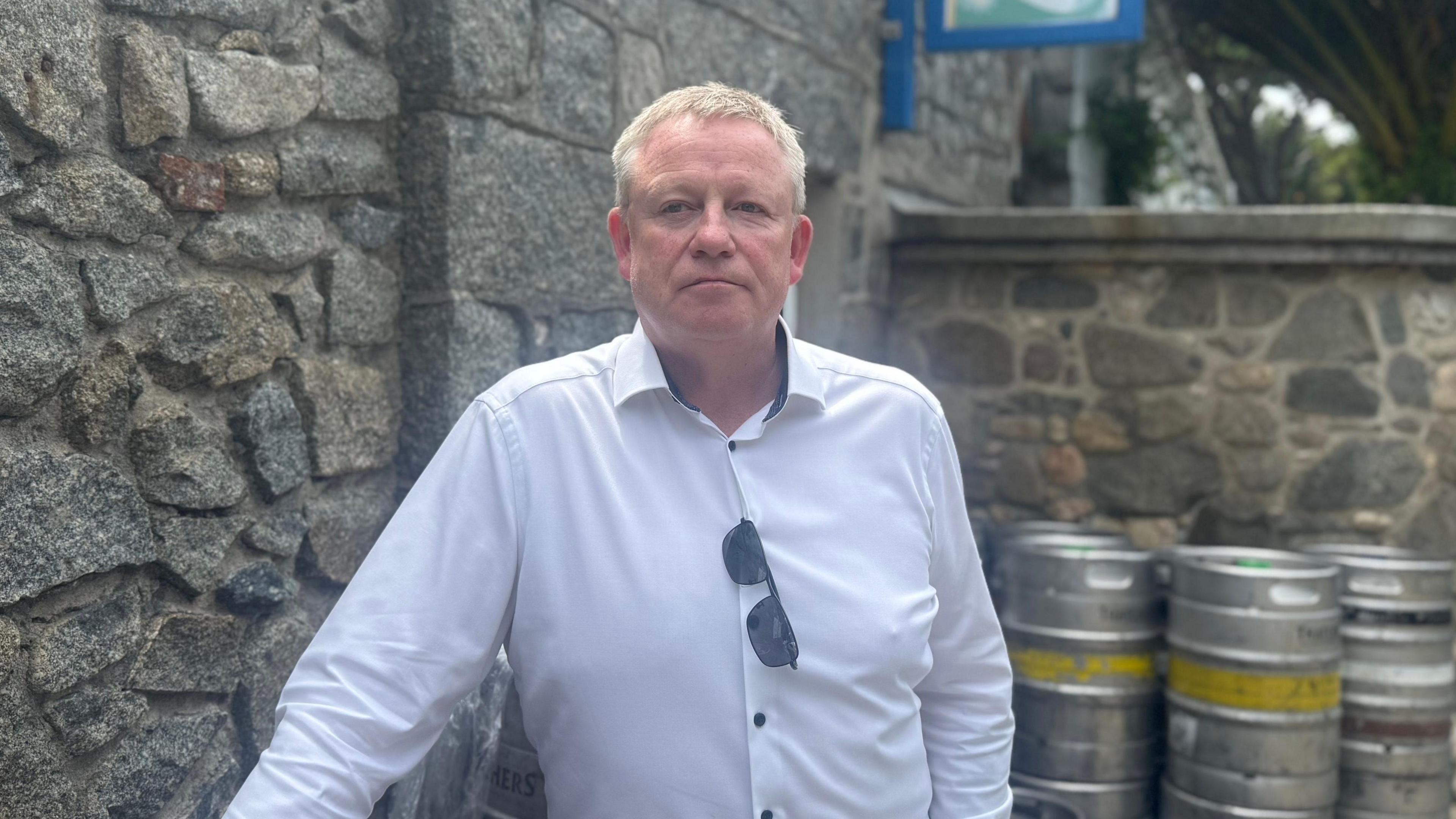 Tom Jones, a fair hared, freckled man in his 50s stood leaning against a bar cannister. He is wearing a white button shirt with black buttons and his sunglasses are hooked on his shirt. He is staring passed the lens.