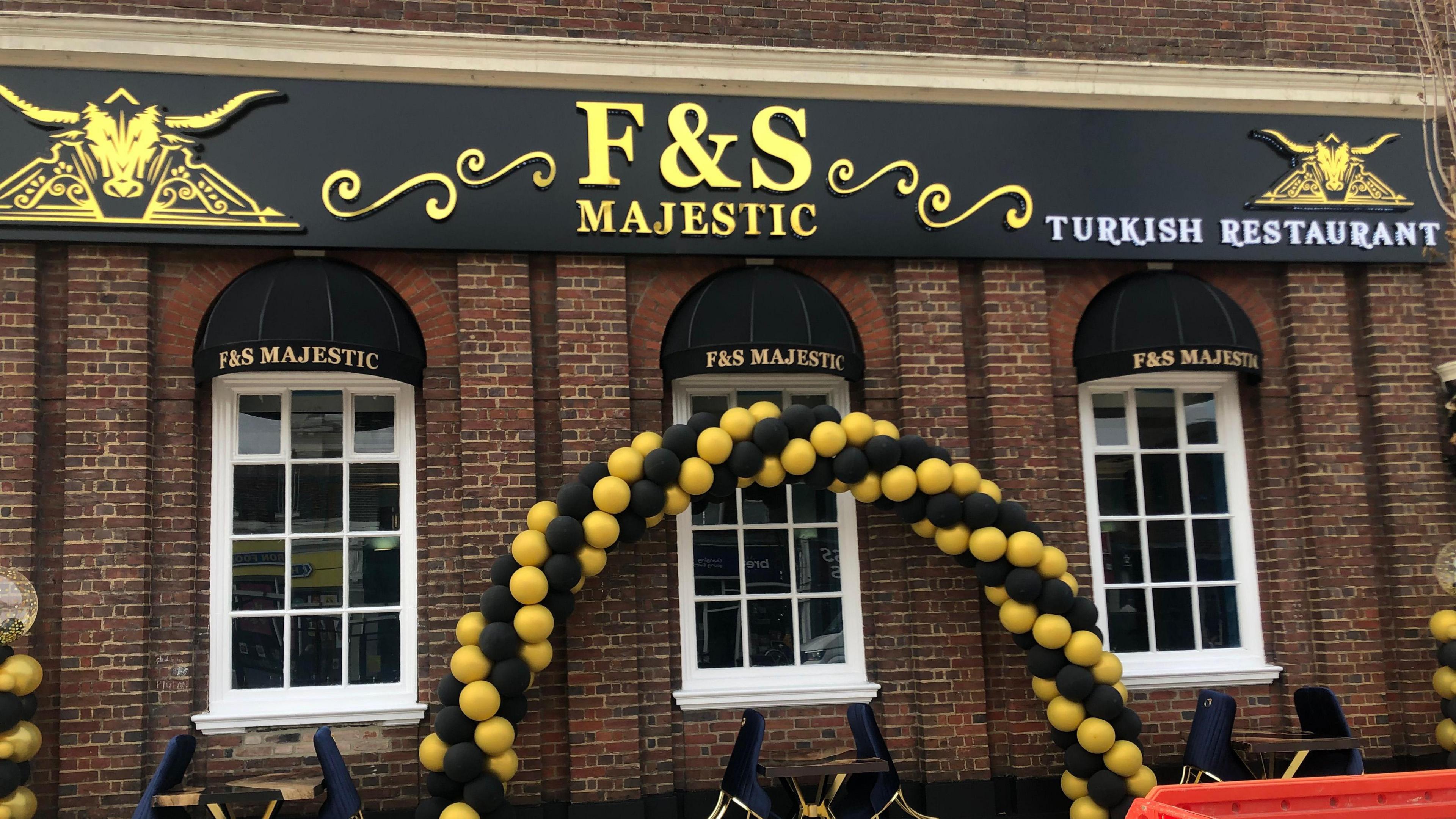 A Georgian style building with three large windows with 12 panes of glass in each one, the windows have black canopies over them with the words 'F&S Majestic' written on them in gold lettering. An arch of black and gold balloons are in the centre of the outside of the building and were used when the establishment opened. Plush tables and chairs can be seen outside on the pavement.