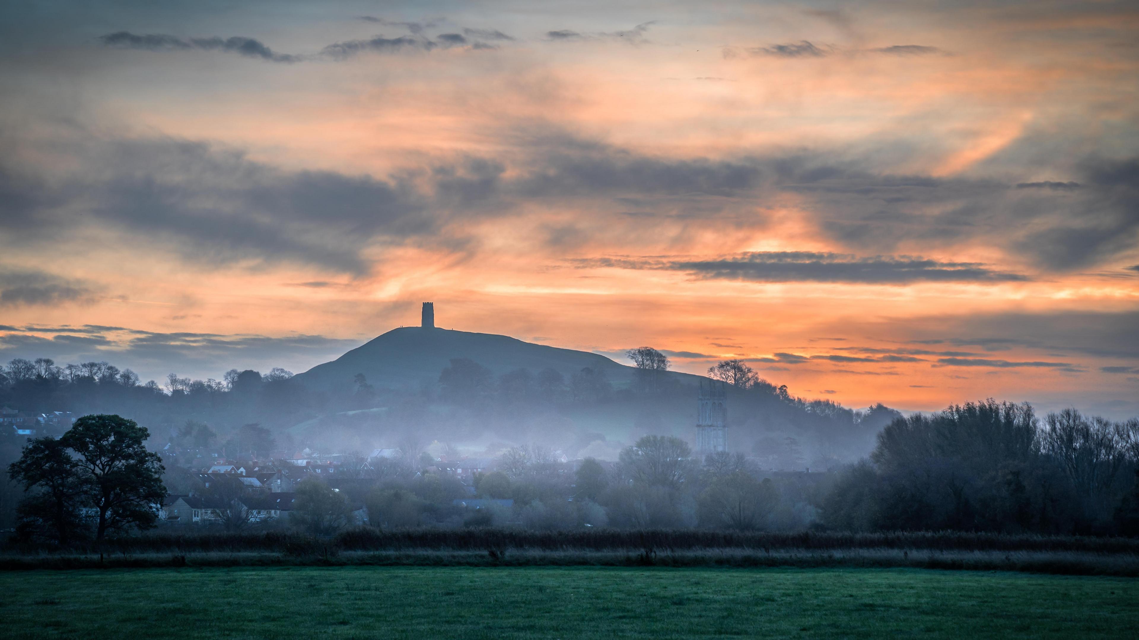 A view of Glastonbury Tor as the sun rises behind it with fog rising from the fields below.