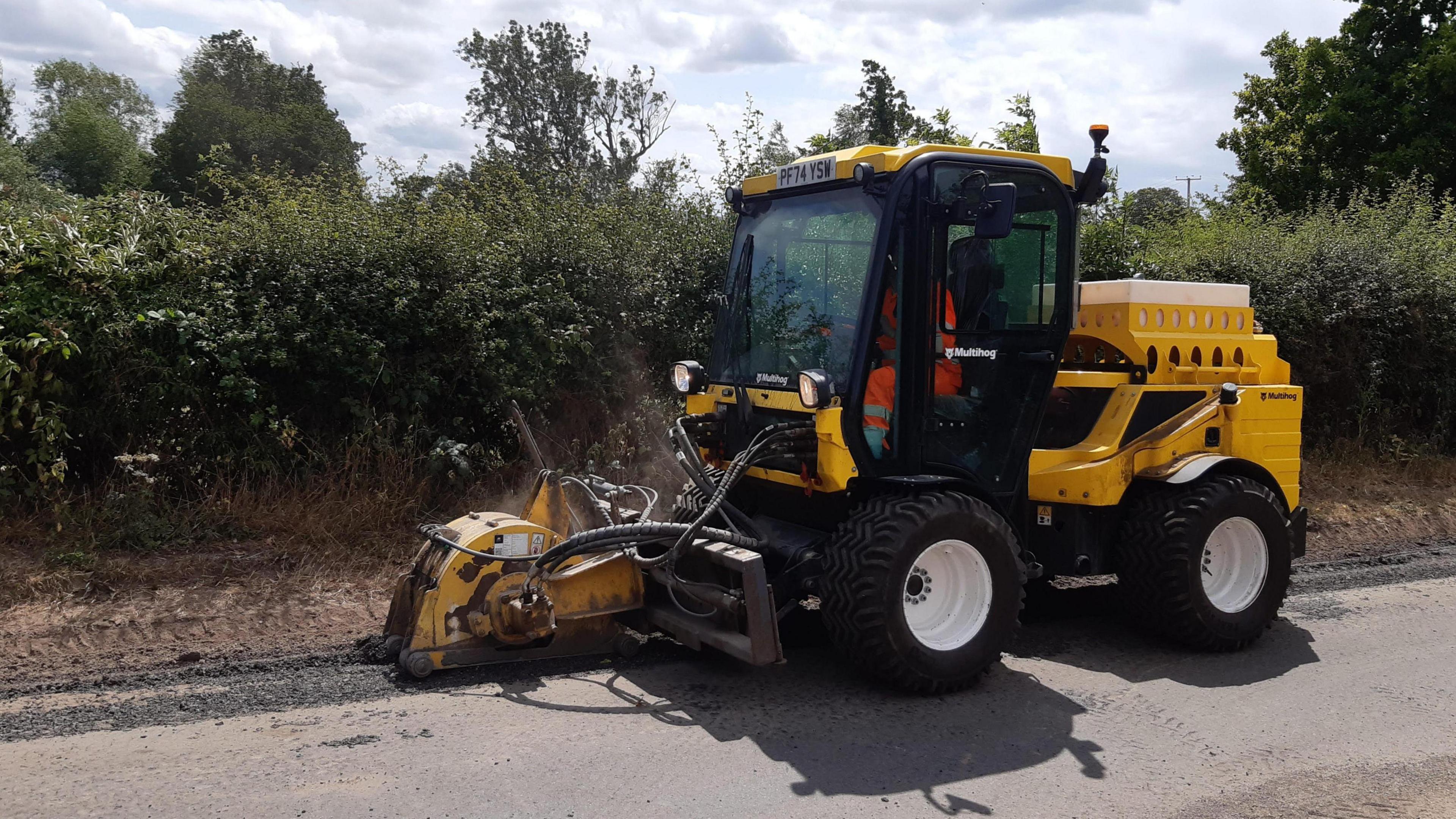 A yellow vehicle with machinery on the front that is filling in a defect on a country road. Inside the vehicle's cab is a person with a hi-vis orange outfit on