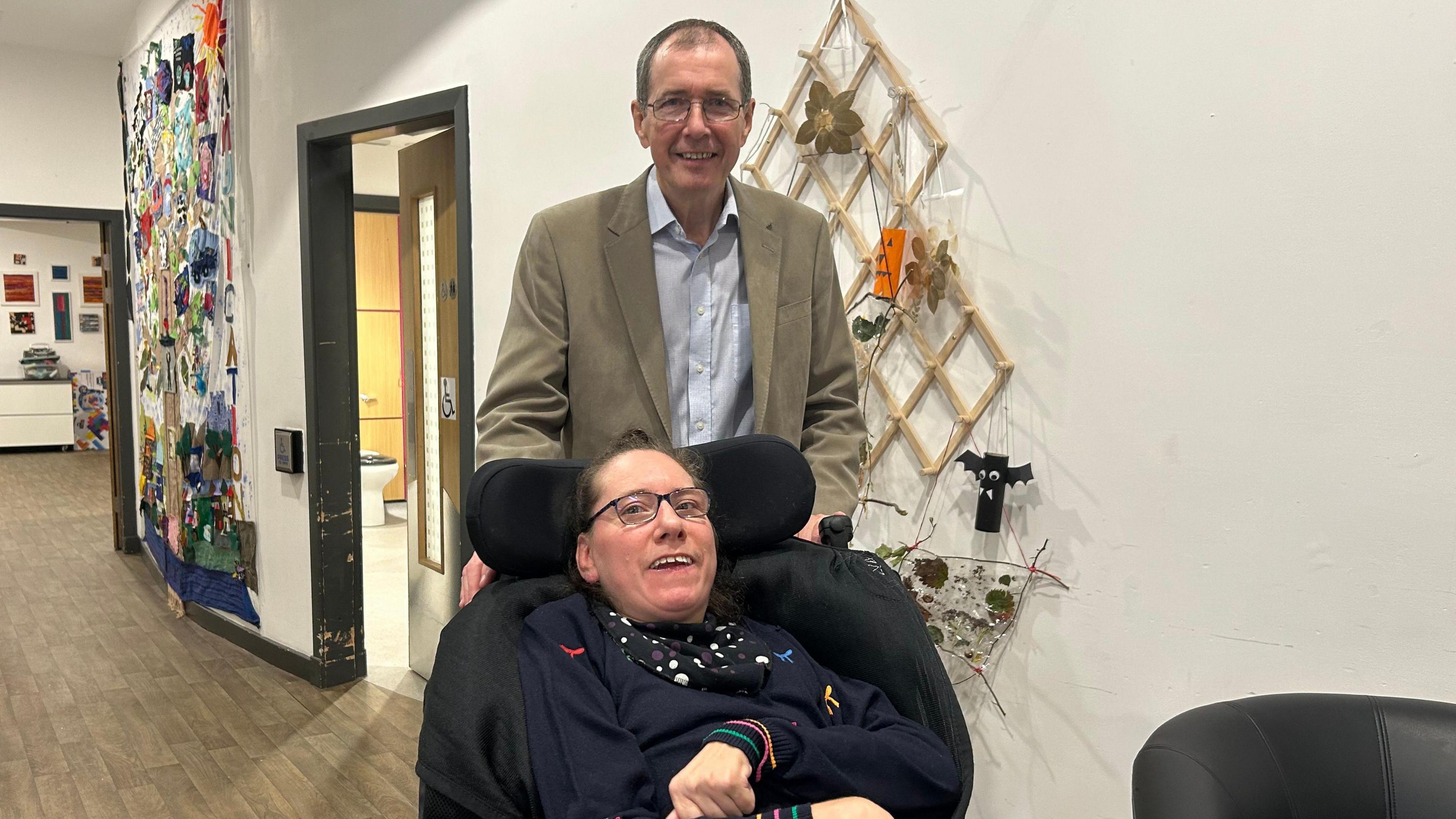 Claire Dempsey, a 48-year-old woman, sits in her wheelchair, she wears a navy jumper with rainbow trim and has glasses. Her father Chris - in a beige jacket and pale blue shirt smiles as he pushes the chair. They appear to be in the corridor of a university building.