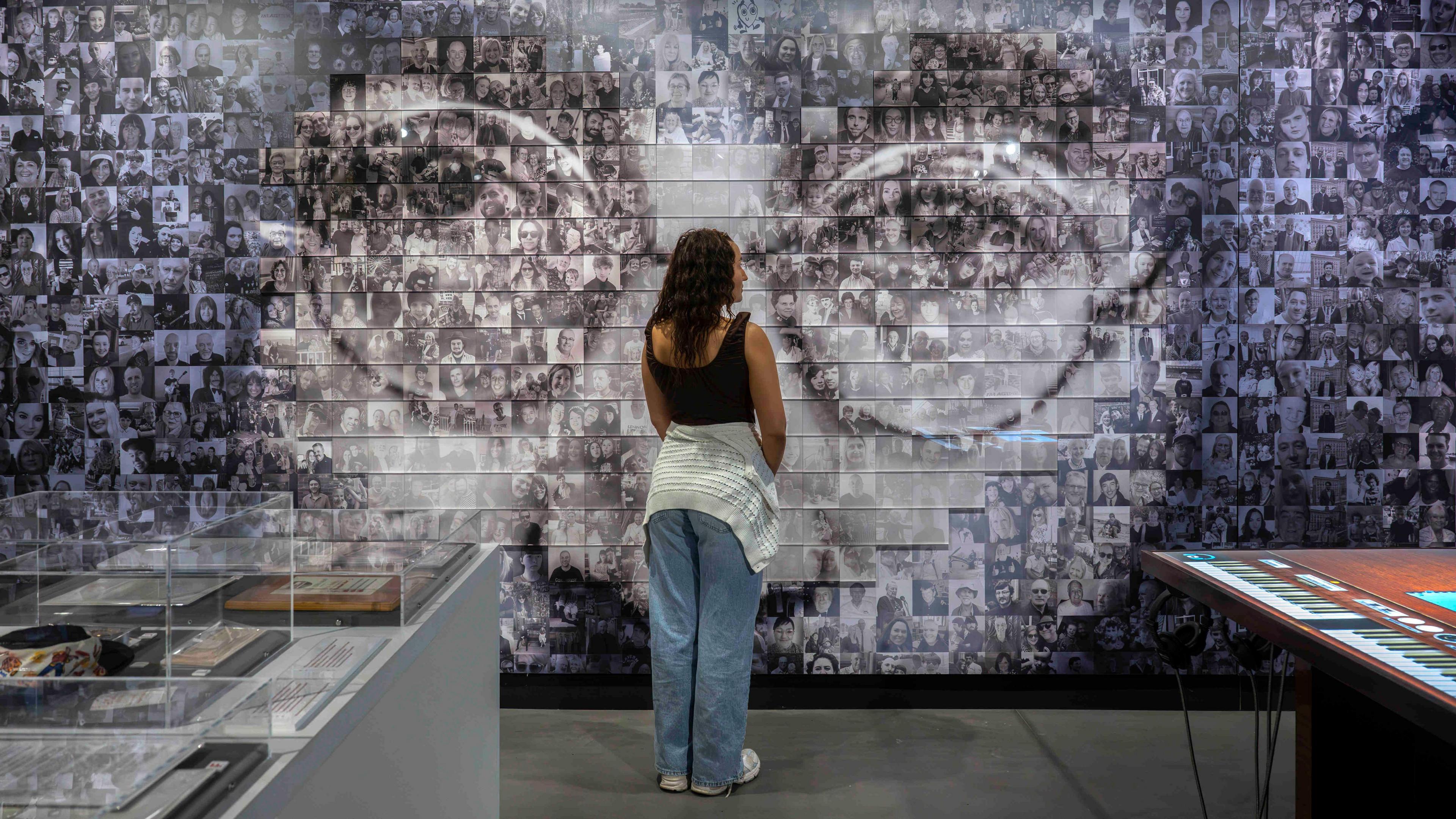 A woman in a black vest and blue jeans stands in front of a black and white photo collage of John Lennon's bespectacled face