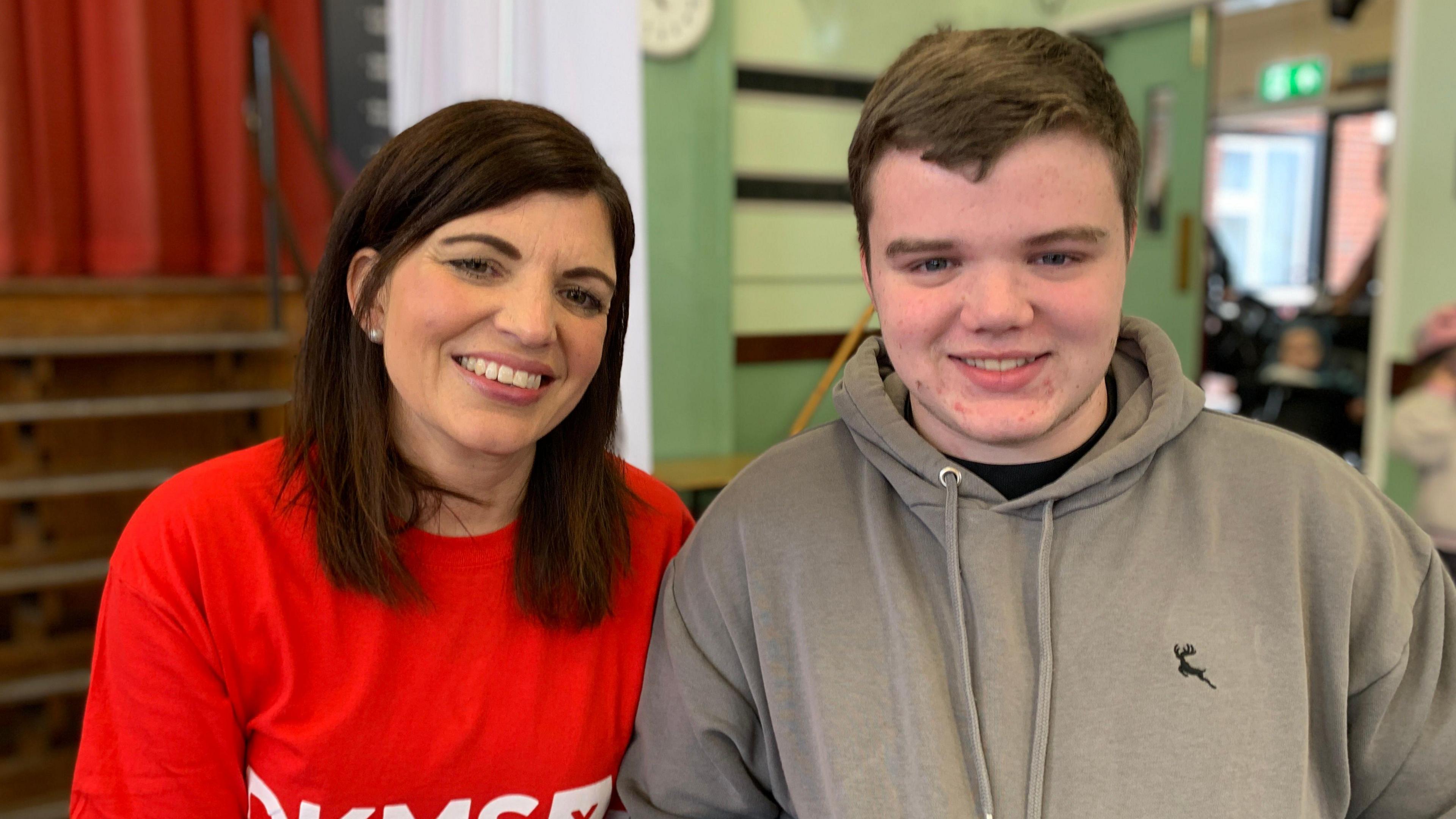 Alana sits with her nephew Robin. She has shoulder-length brown hair and is wearing a red T-shirt. He has short brown hair and is wearing a grey hoody with a logo of a leaping mouse in on the left breast.